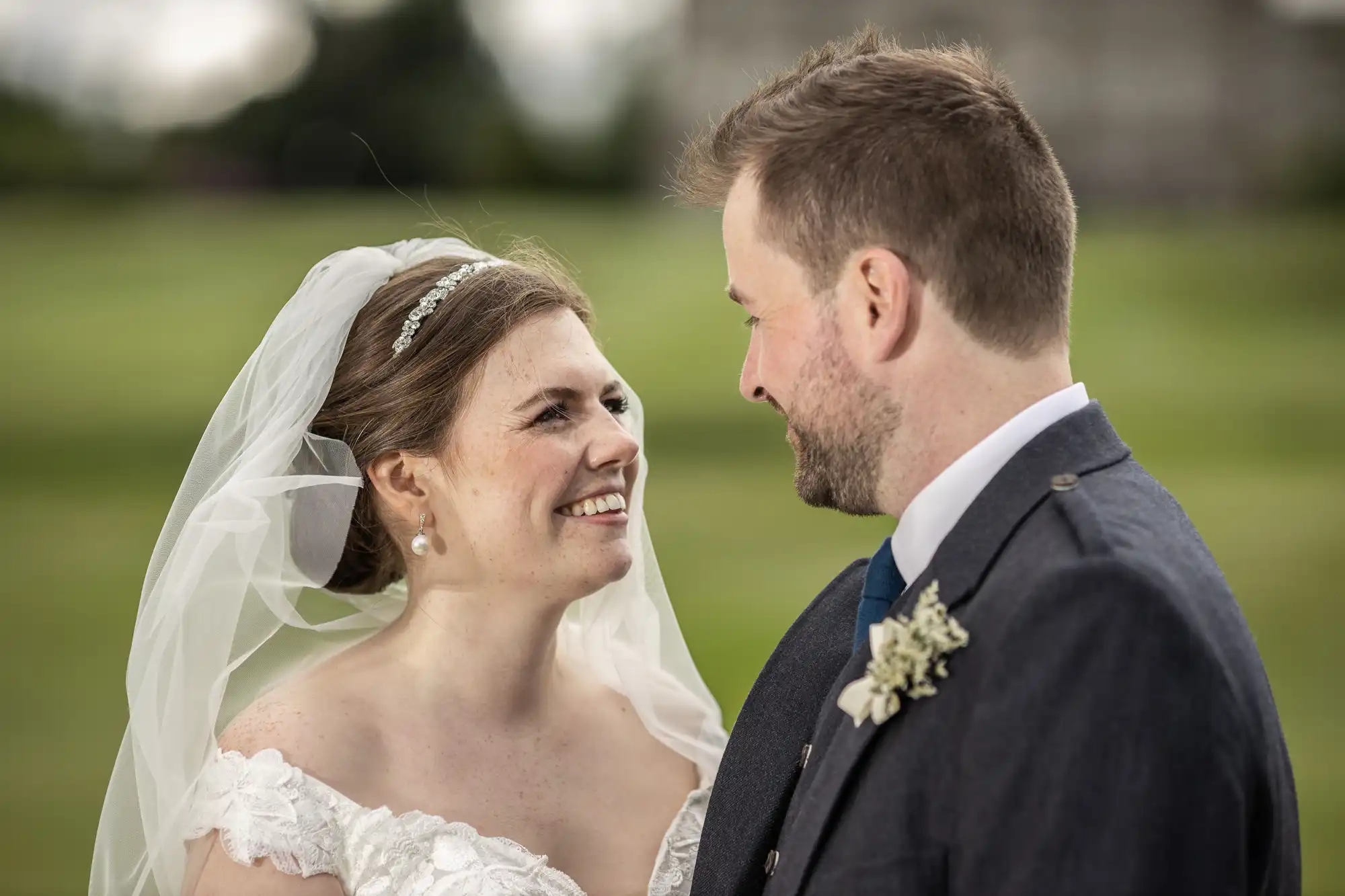 A bride and groom, both smiling, stand facing each other outdoors. The bride wears a white gown and veil, while the groom is in a formal suit with a boutonnière.