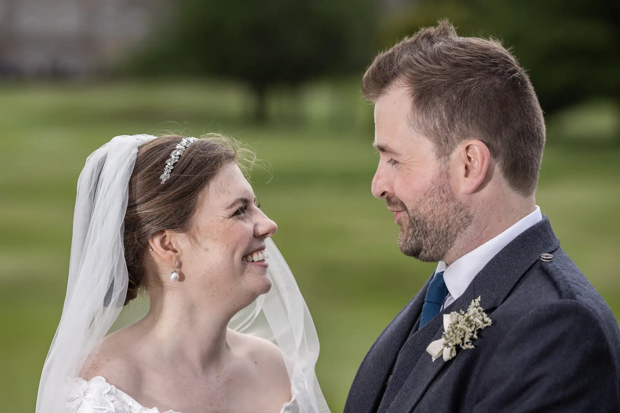 A bride and groom stand outdoors, facing each other and smiling. The bride wears a white dress and veil, while the groom is in a dark suit with a boutonnière.