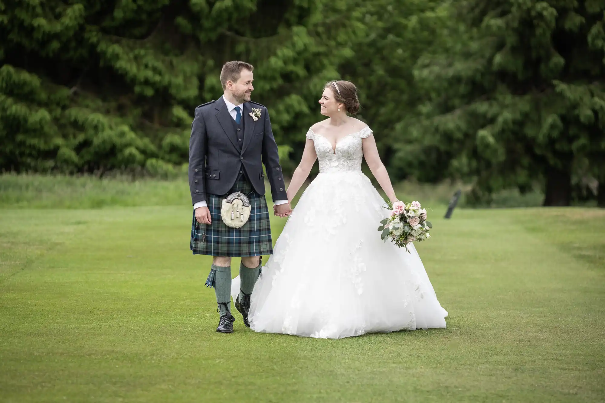 A bride and groom, holding hands, walk on a grassy field. The groom wears a kilt, and the bride is in a white gown, carrying a bouquet. Trees are visible in the background.