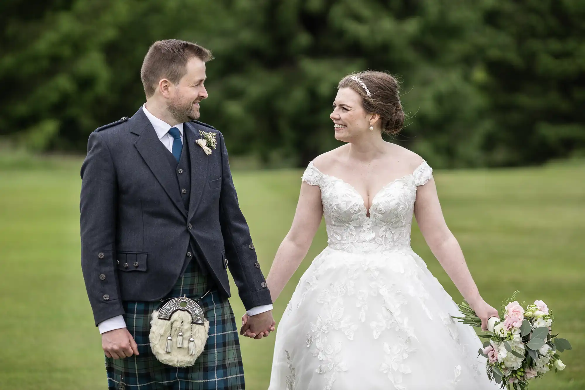 A bride in a white gown and a groom in a kilt and jacket are holding hands and smiling at each other, standing on a green lawn with trees in the background.