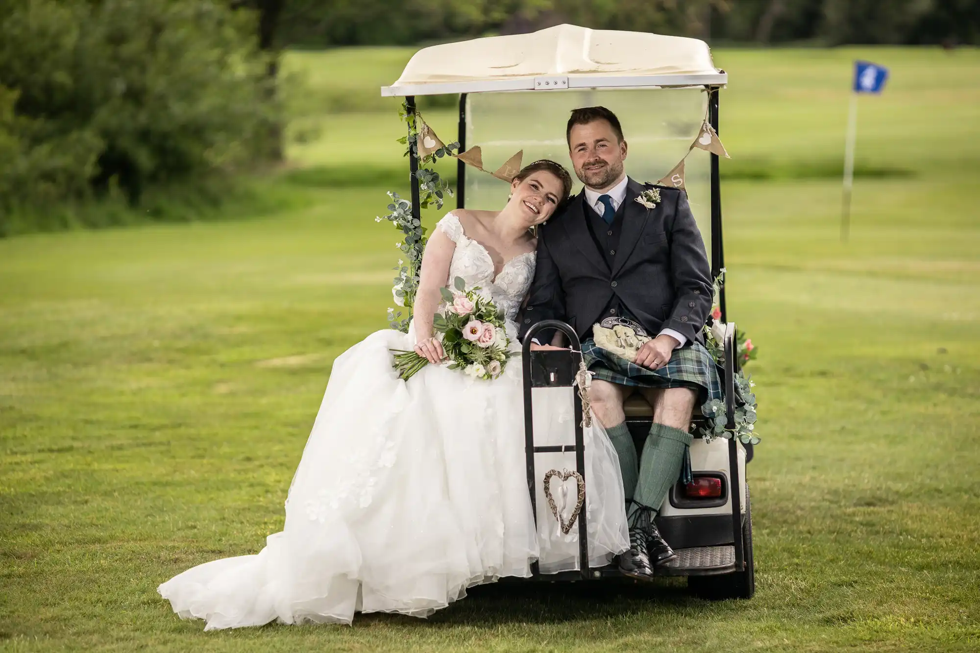 A bride and groom sit on a decorated golf cart on a golf course. The bride leans on the groom's shoulder, holding a bouquet. The groom wears a kilt, and both are smiling.