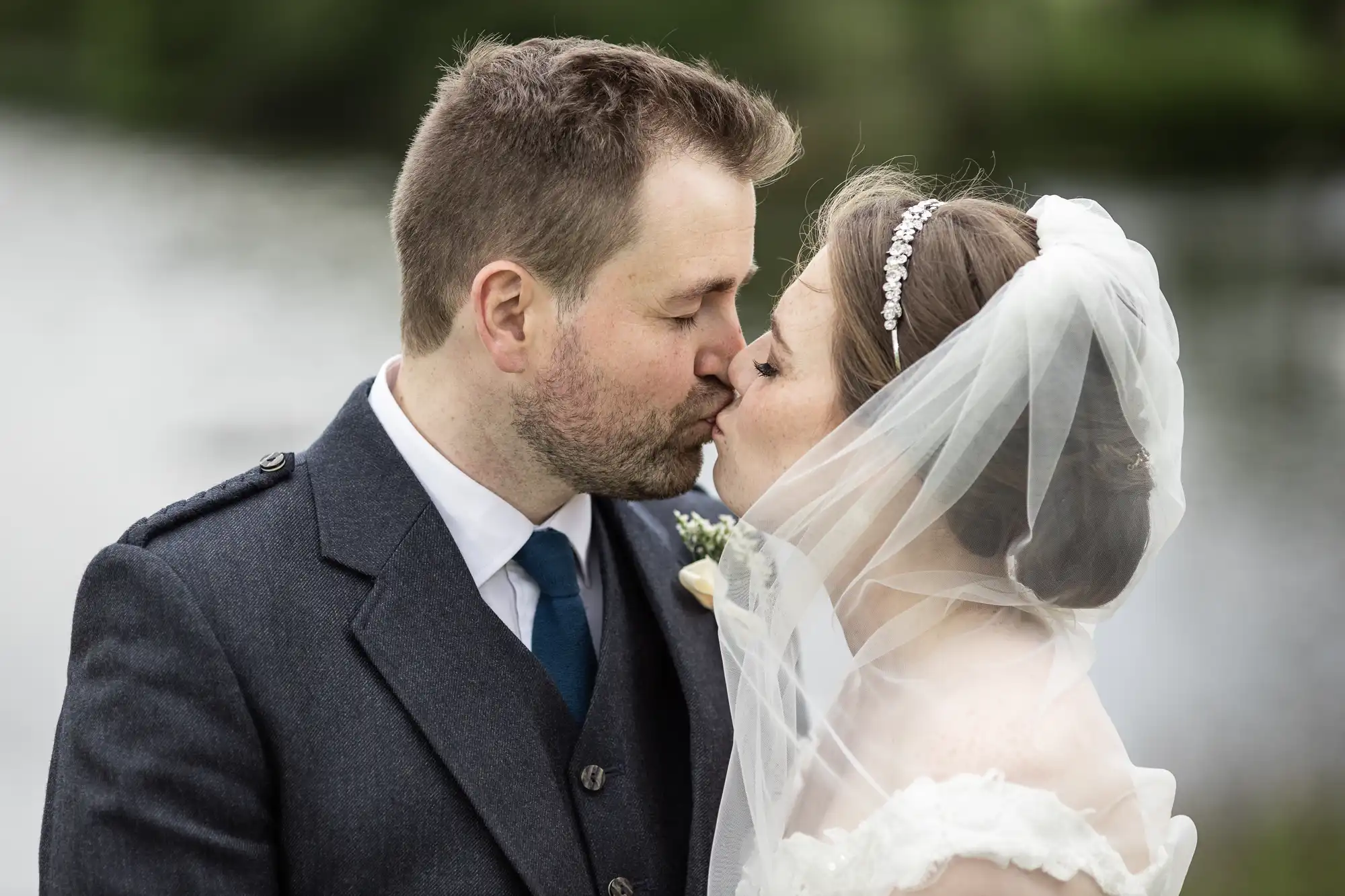 A bride and groom sharing a kiss outdoors, the bride wearing a veil and headband, and the groom in a dark suit and tie.