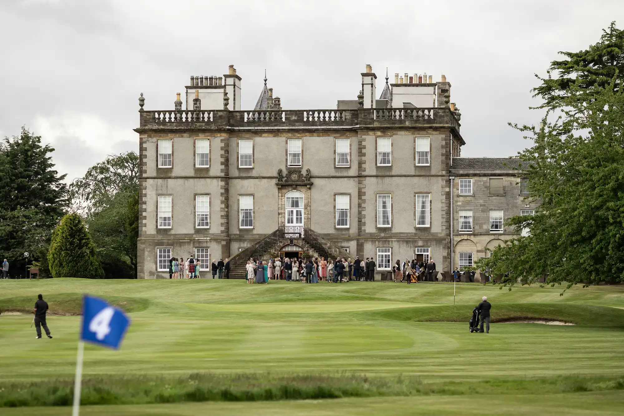 A large group of people gather outside a historic mansion with manicured lawns and a blue golf flag numbered 4 in the foreground.