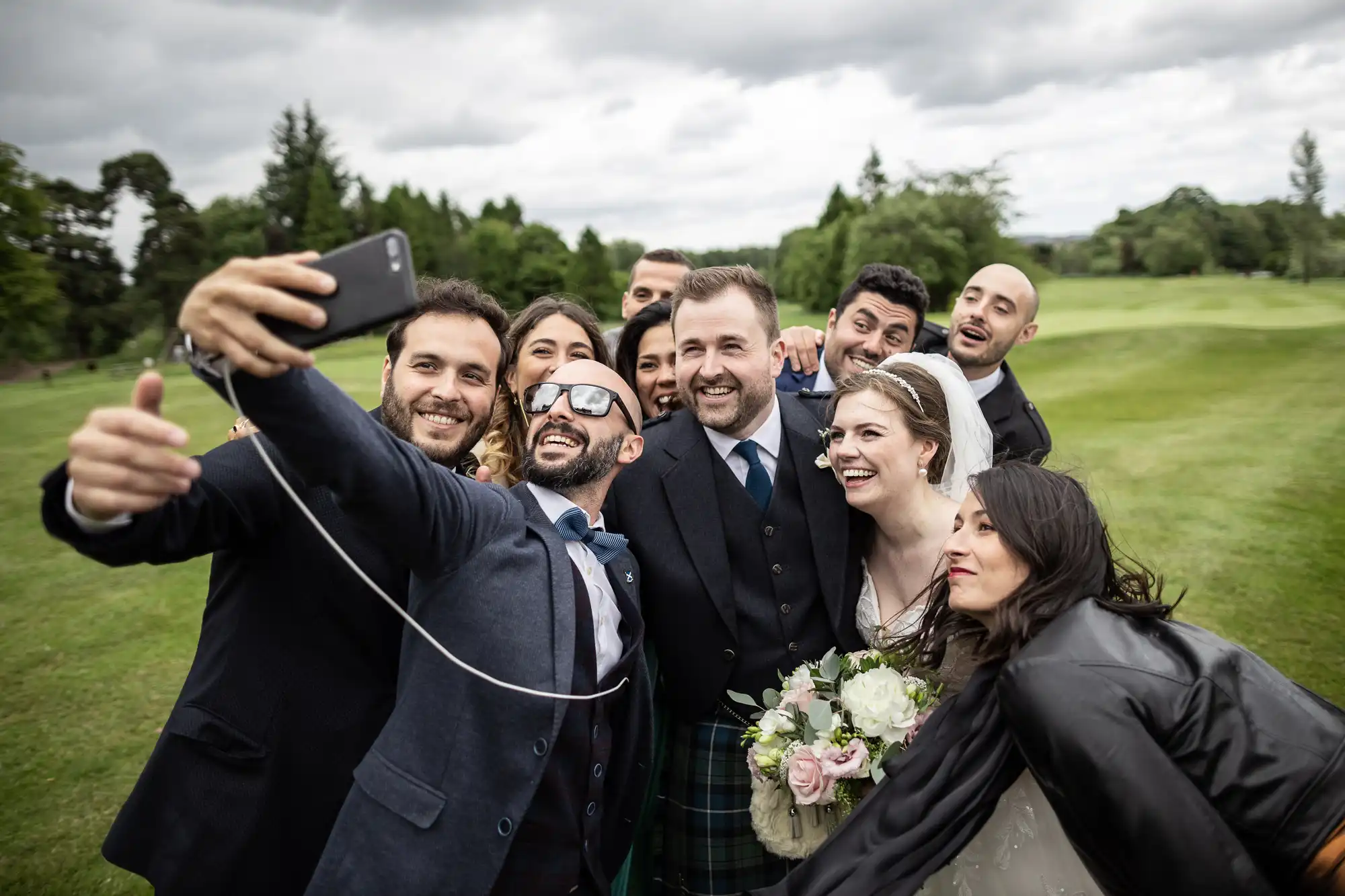 A group of people gathered outdoors, smiling and taking a selfie. The individual holding the phone is in the foreground, and a bride in a white dress and veil is also visible in the crowd.