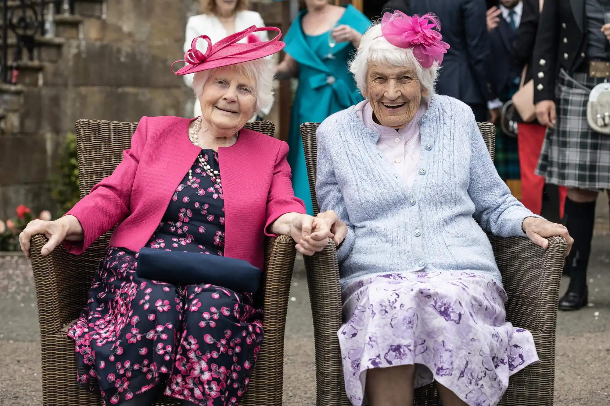 Two elderly women, dressed in vibrant outfits and hats, sit side by side in wicker chairs, holding hands and smiling. They are outdoors, with others blurred in the background.