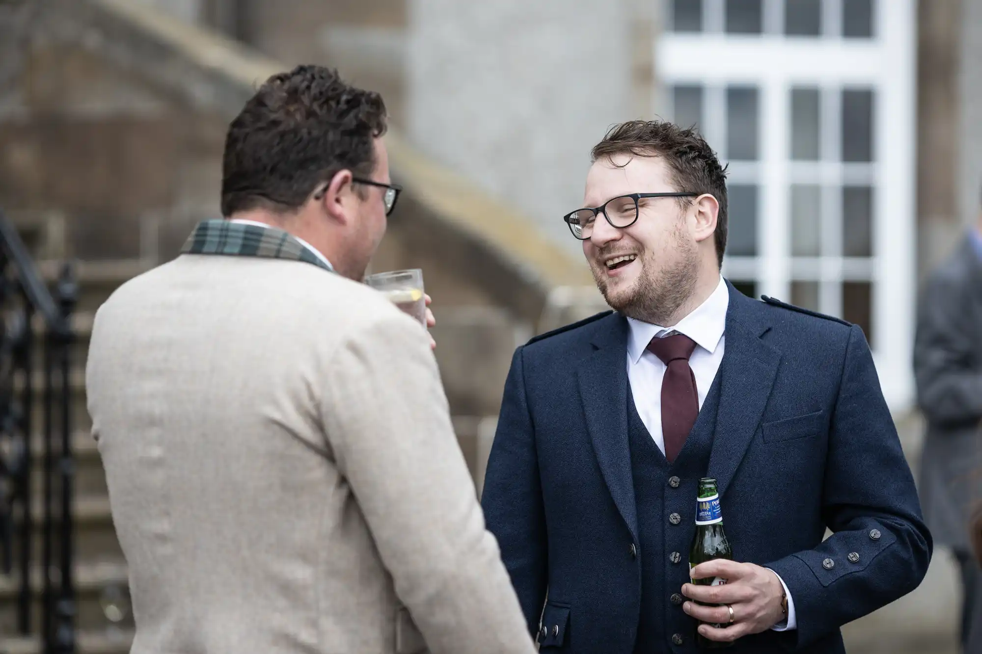 Two men in suits converse and laugh while holding drinks, one with a beer bottle and the other with a glass, standing outside a building with a staircase and a window in the background.