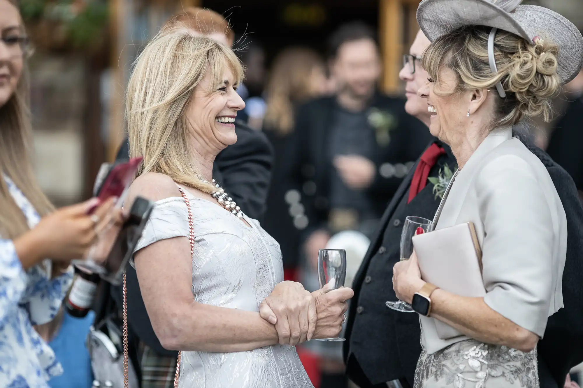 Two women in formal attire share a laugh at an outdoor social event, both holding drinks. Other people are in the background, and one woman is holding a smartphone with a red cover.