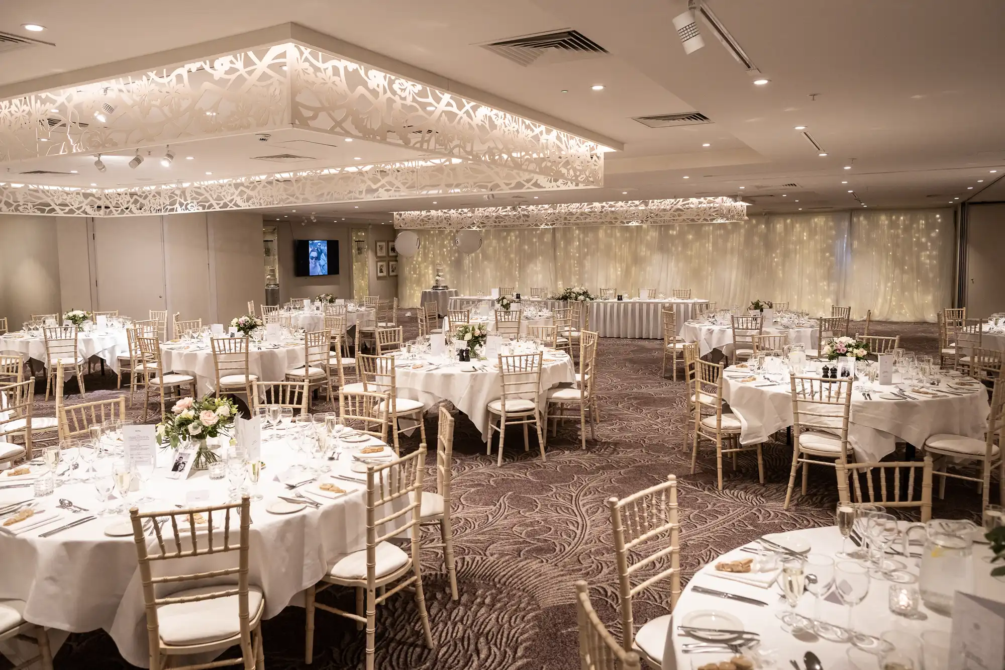 A large, elegantly decorated banquet hall with round tables set for dining, adorned with white tablecloths, floral centerpieces, and surrounded by wooden chairs. A long table is situated at the far end.