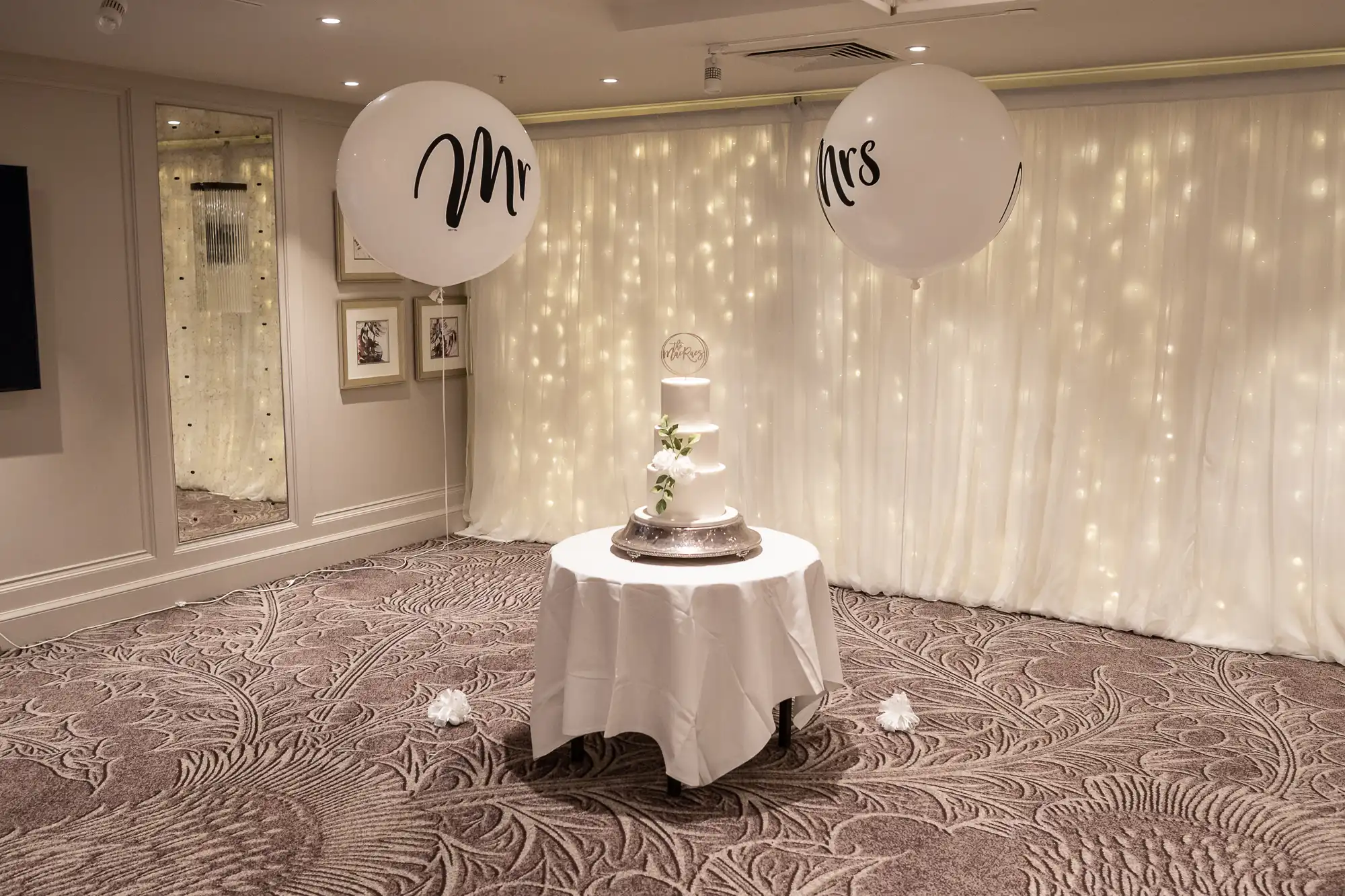 A white frosted wedding cake on a round table with "Mr" and "Mrs" balloons, set against a backdrop of twinkling lights in an elegantly decorated room.