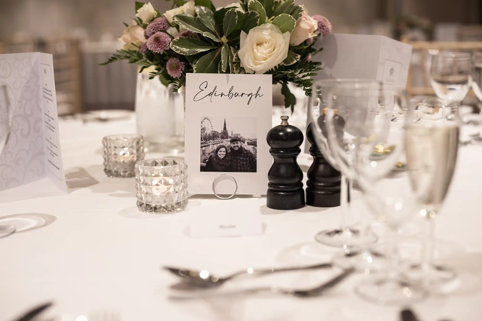 A dining table set with wine glasses, cutlery, candle holders, and a floral centerpiece with a card labeled "Edinburgh" featuring a photo of two people and the Edinburgh skyline.