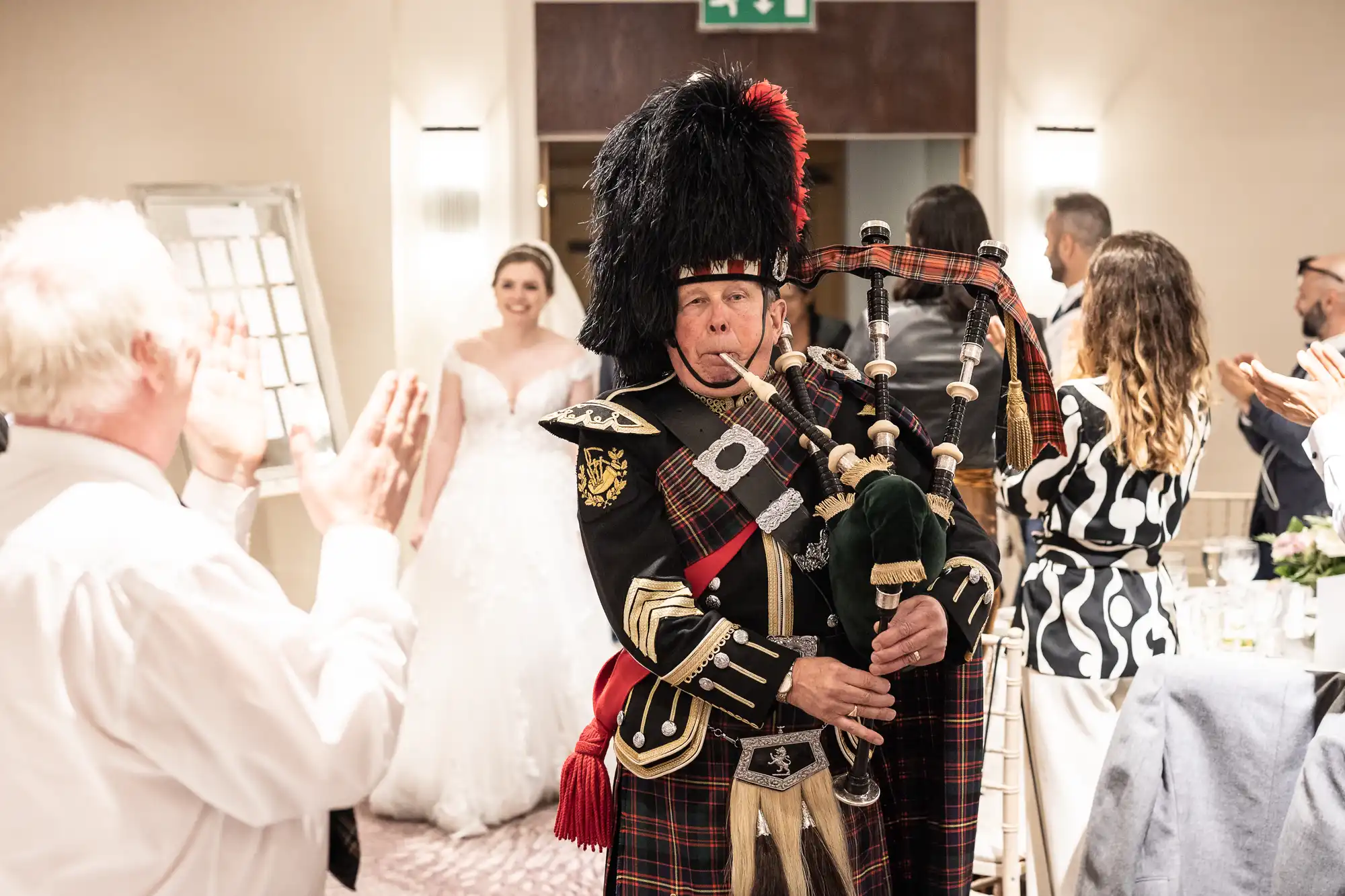 A bagpiper in traditional Scottish attire plays the bagpipes in a room with people clapping and a bride in a white gown smiling in the background.