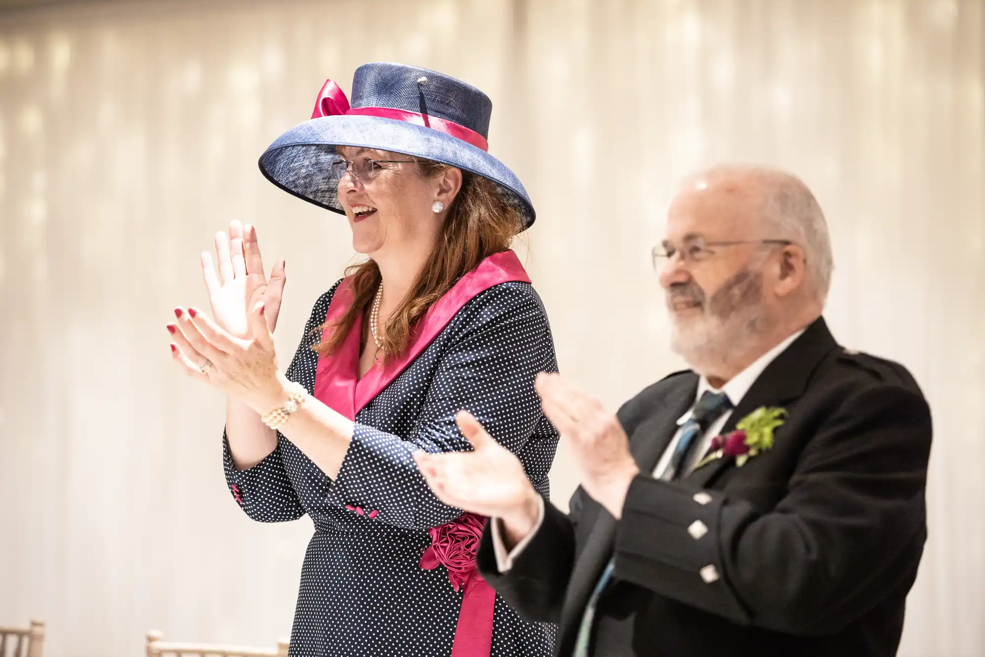 A woman in a hat and a man in formal attire are standing and clapping at an event.