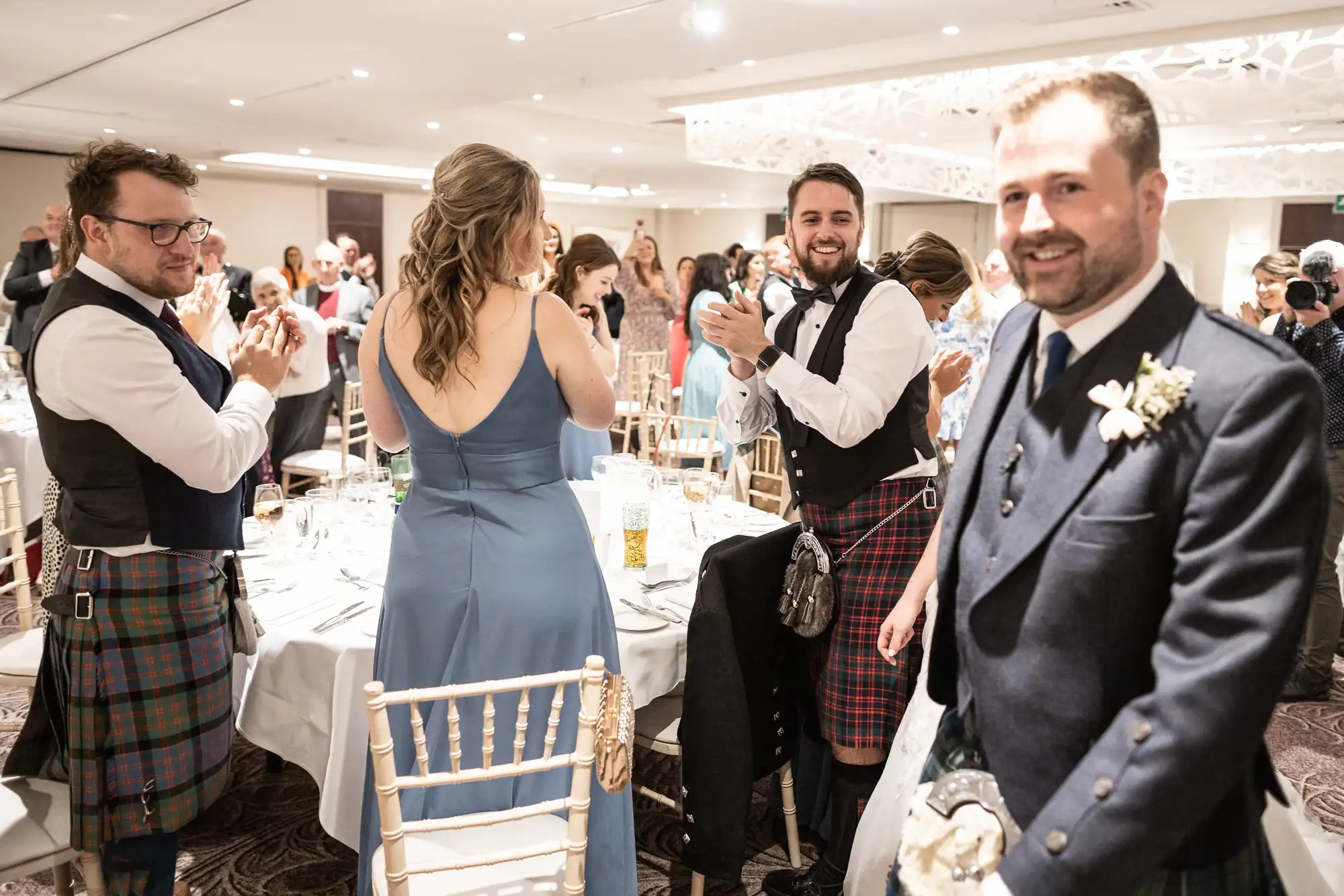 People in formal attire, some wearing kilts, stand and applaud around a set dining table in a bright, elegant room.