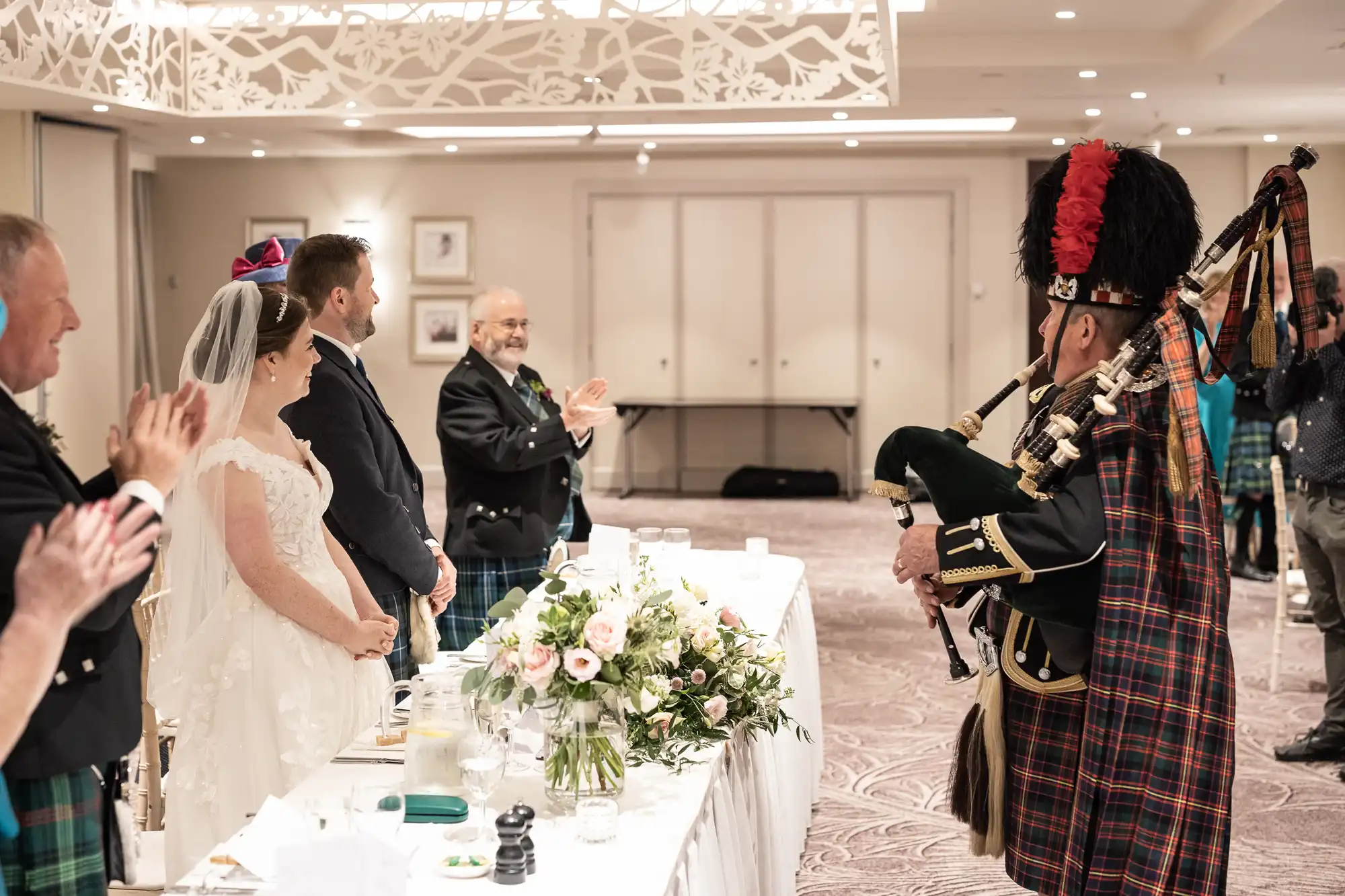 A bride and groom in wedding attire stand at a table adorned with flowers. Guests applaud as a bagpiper dressed in traditional Scottish attire plays the bagpipes in front of them.