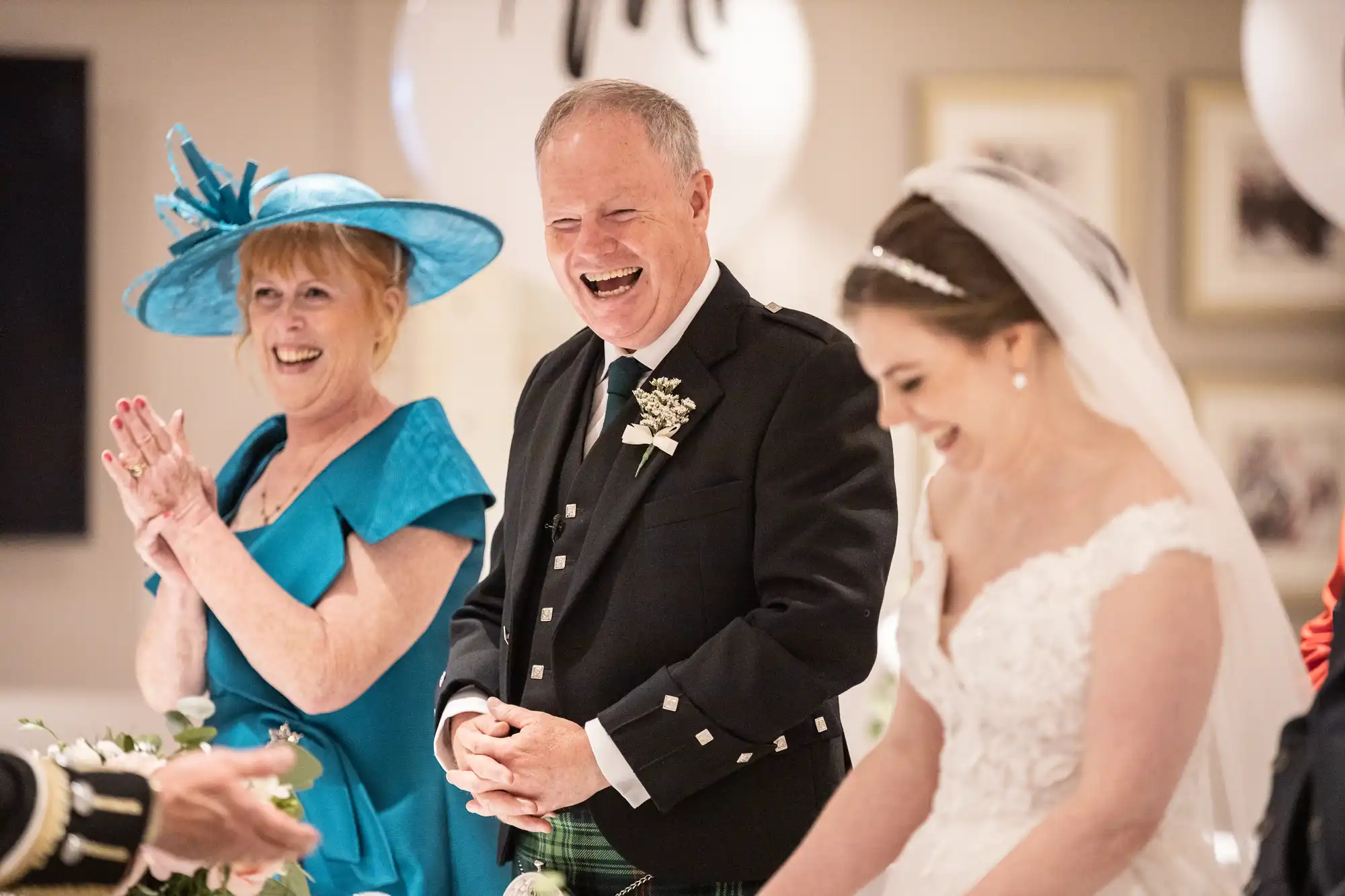 A man in a kilt, a woman in a blue dress and hat, and a bride in a white gown share a moment of laughter at a wedding.