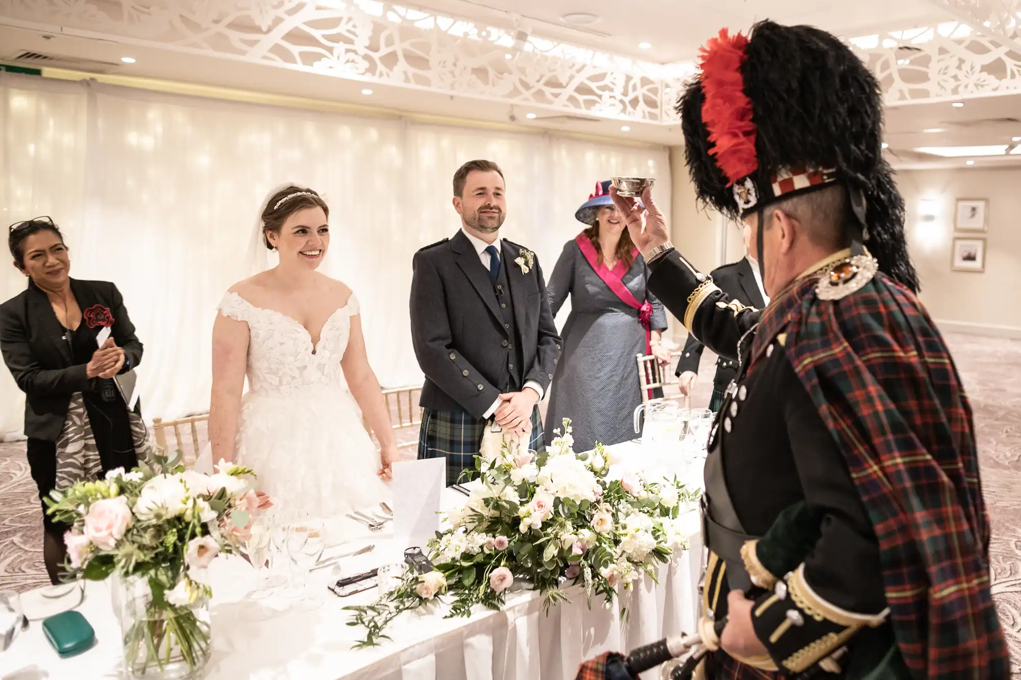 A bride and groom, alongside well-dressed guests, stand at a decorated table while a man in traditional Scottish attire raises a toast.