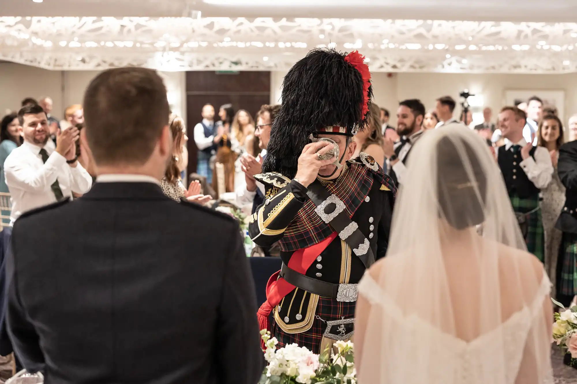 A man in traditional Scottish attire, playing bagpipes, is facing a bride and groom in a crowded reception hall with guests clapping and cheering in the background.