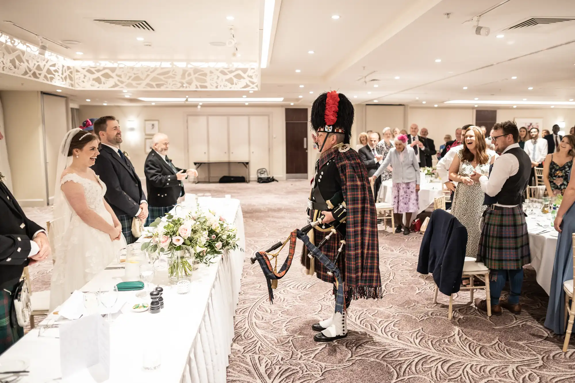 A traditional Scottish wedding ceremony: An individual in full traditional Scottish attire stands before a wedding couple, while guests watch and stand in an elegantly decorated hall.