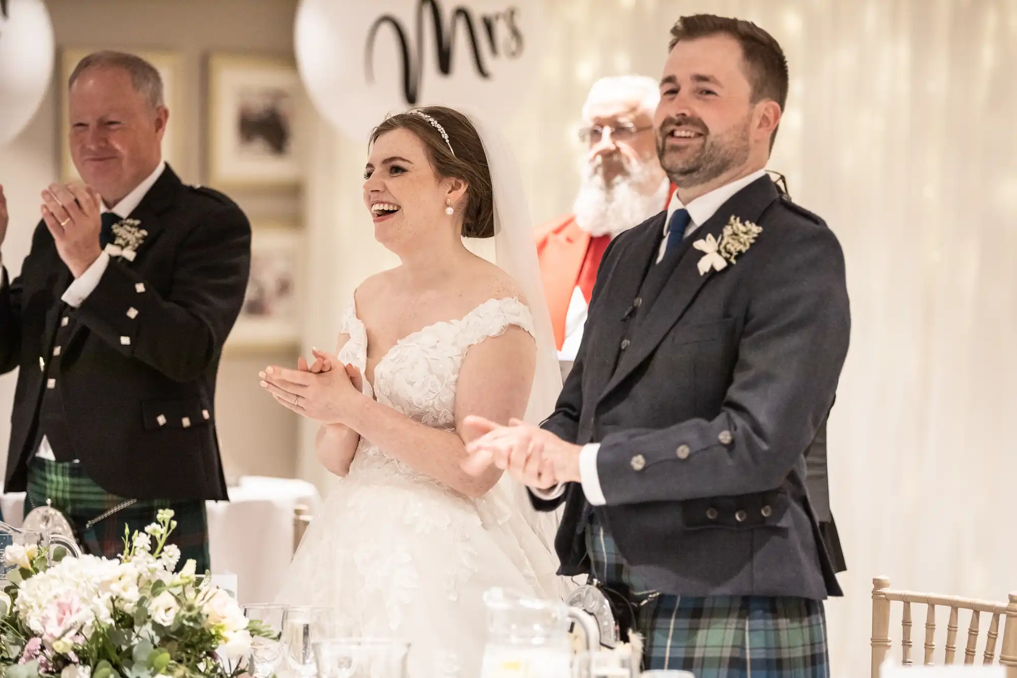 Bride and groom in wedding attire, smiling and clapping at a reception. Two men, also in formal attire, stand beside them, clapping. A "Mrs" decoration is visible in the background.