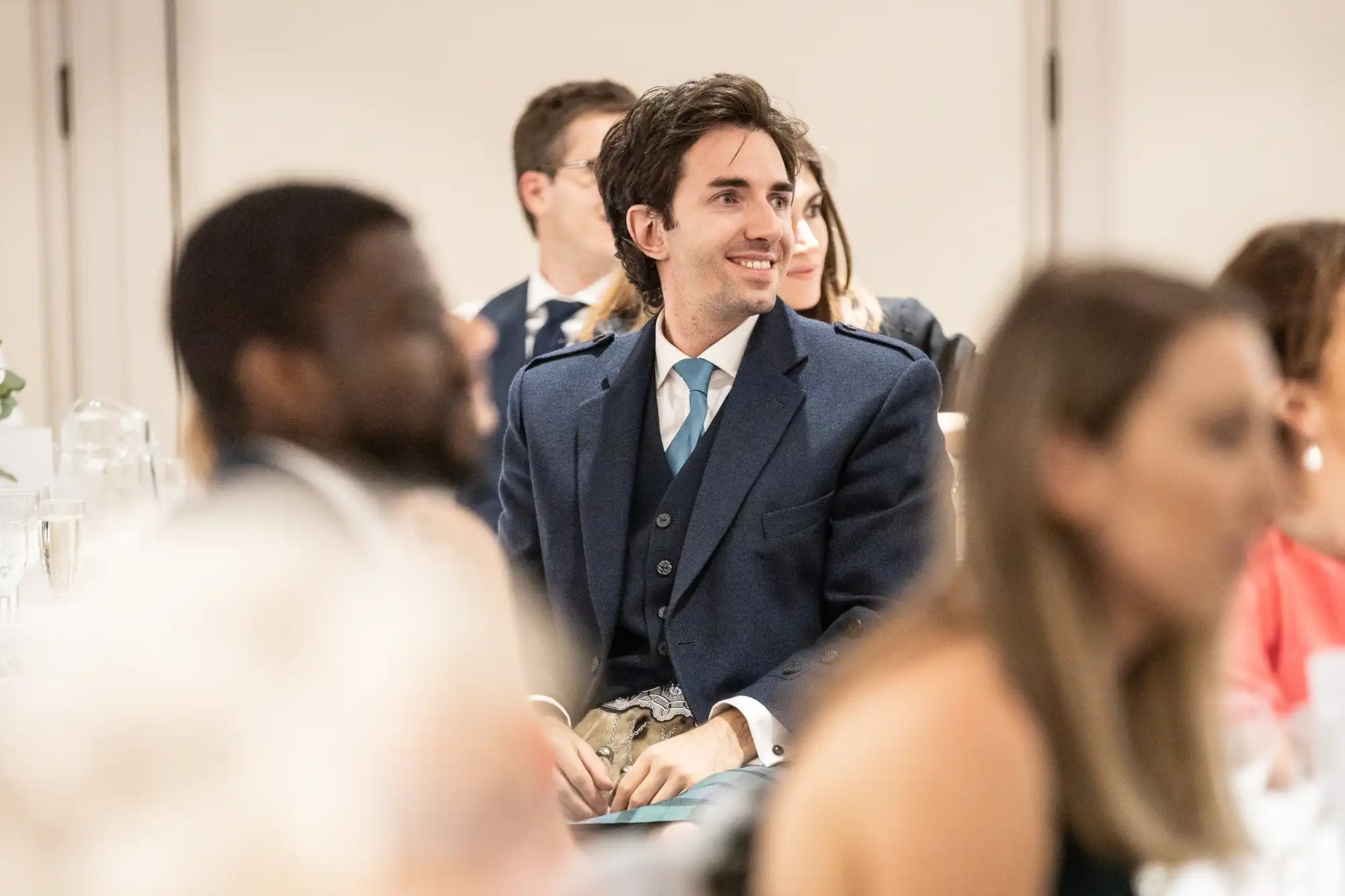 A man in a suit and tie sits at a formal event, smiling and looking to the side. Several people are seated around him, focused on the event.