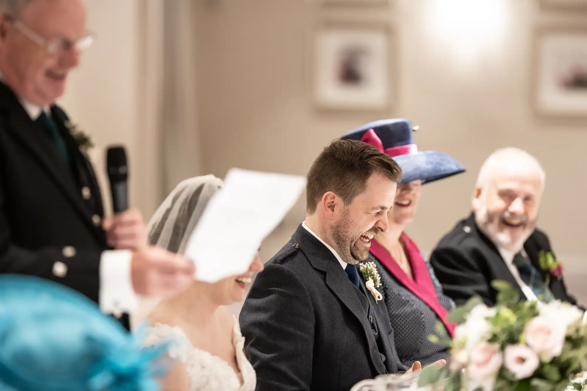 A wedding reception scene with five people laughing and listening to a speech. The bride and groom are seated at a table adorned with flowers, while three guests in formal attire are nearby.