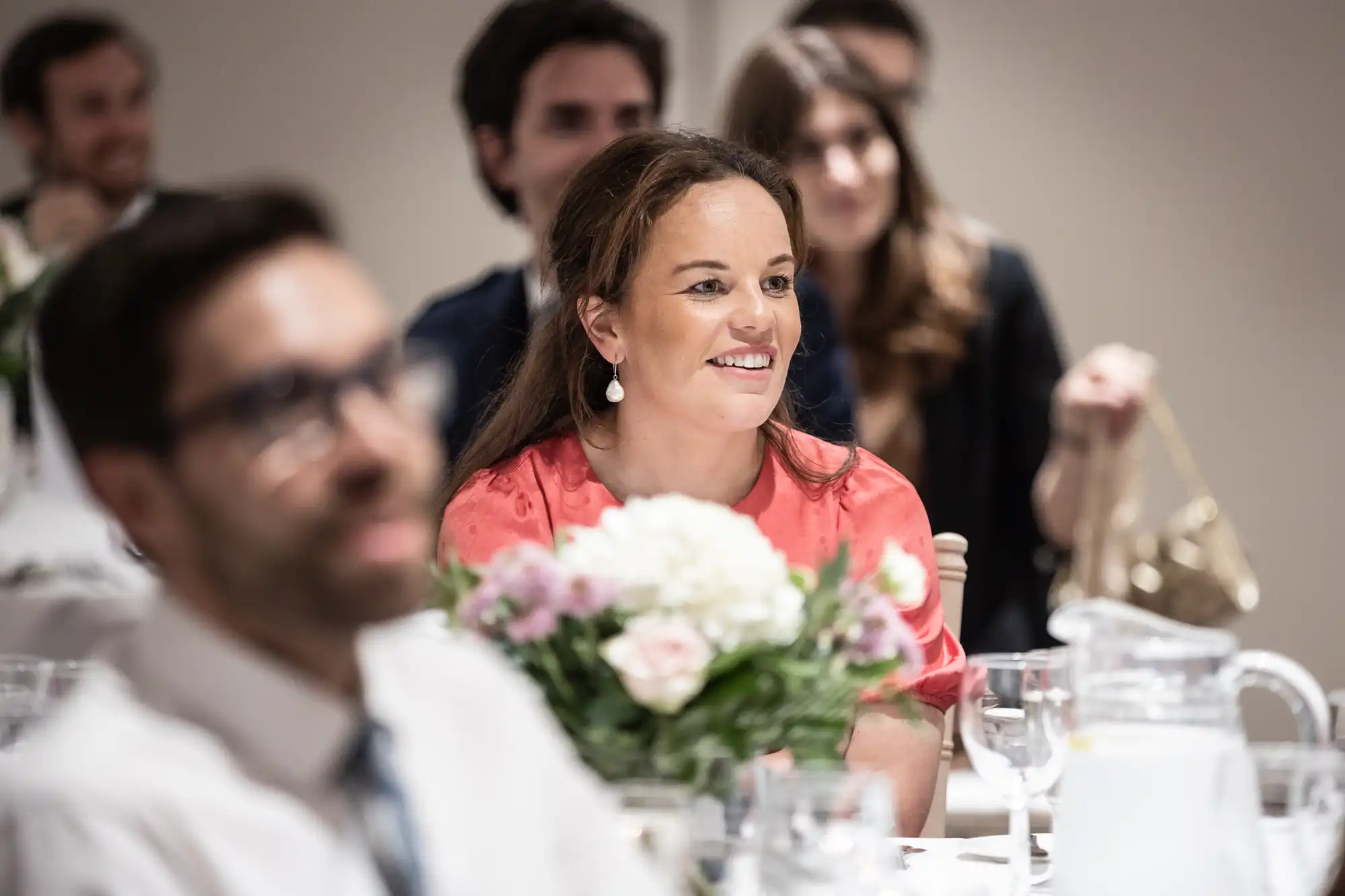 A woman in a red top attentively listens during an event, surrounded by others seated at tables with floral centerpieces and glassware.