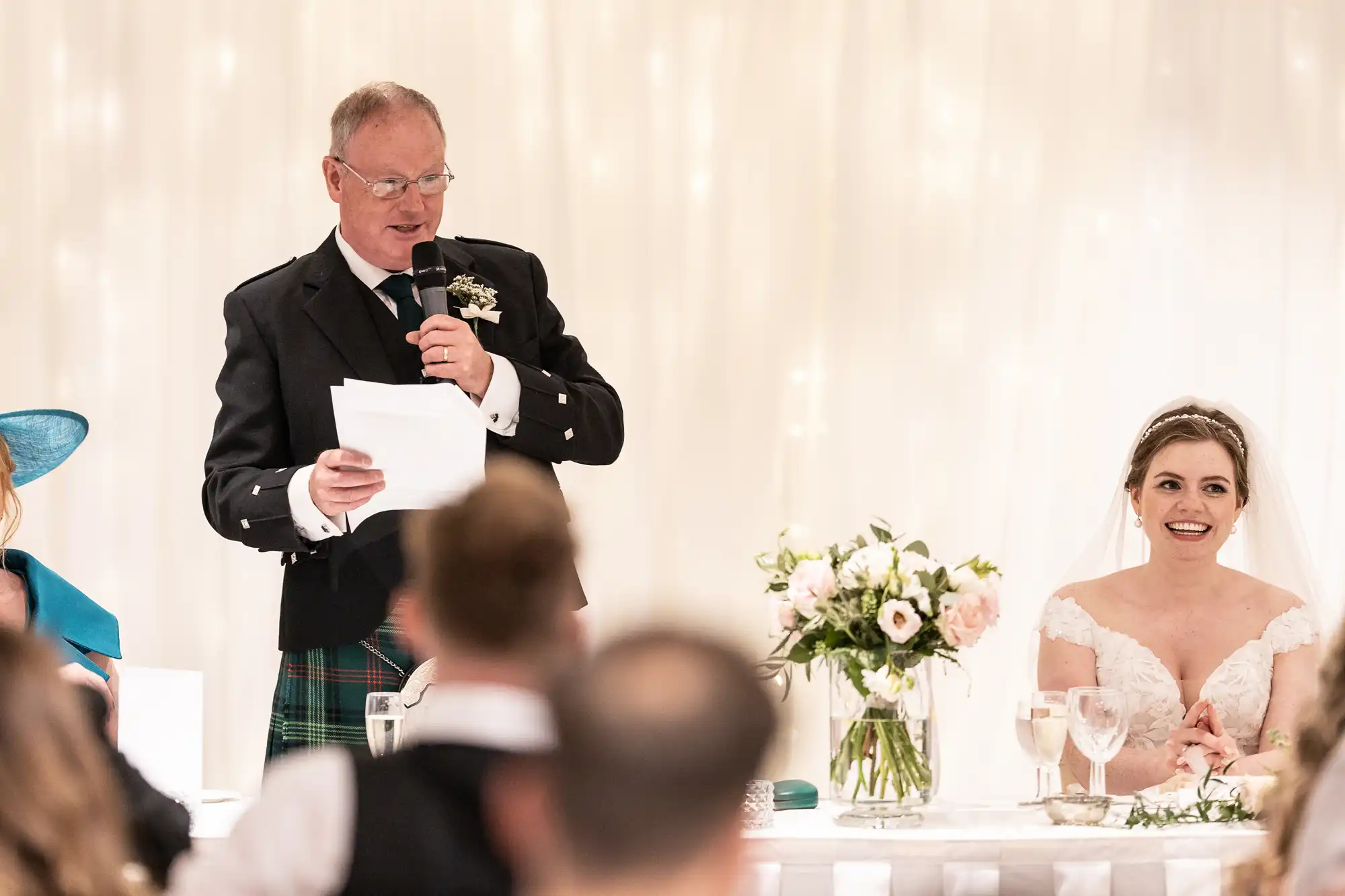 A man in a traditional outfit gives a speech with a microphone at a wedding reception. A bride in a white dress sits next to him, smiling.