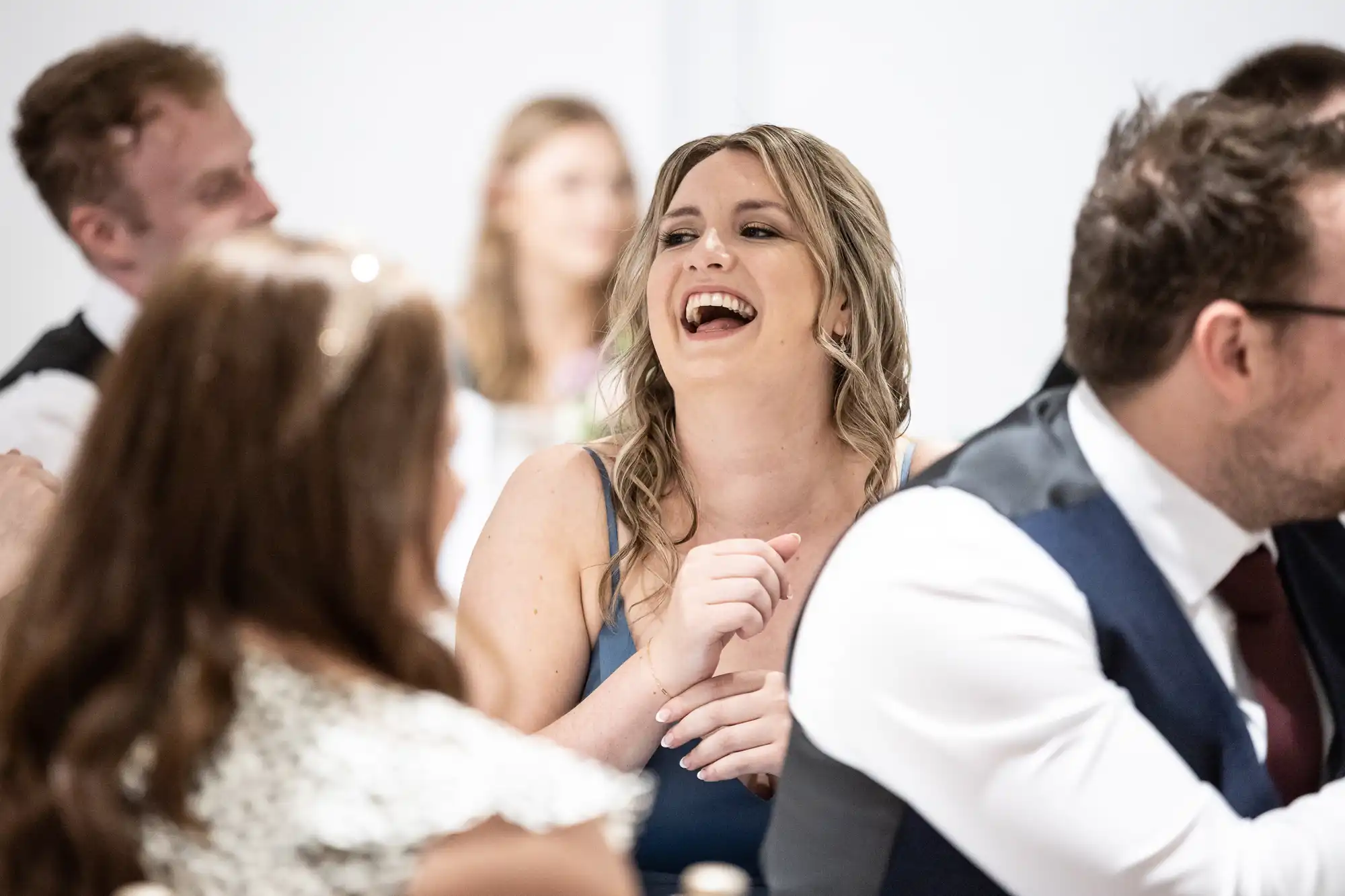 A woman in a blue dress laughs while sitting at a table with other people dressed formally.