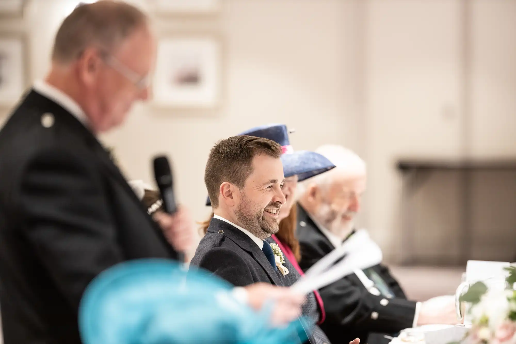 A man in formal attire speaks into a microphone at a table, while another man smiles and listens. Others are seated at the table, some wearing hats.