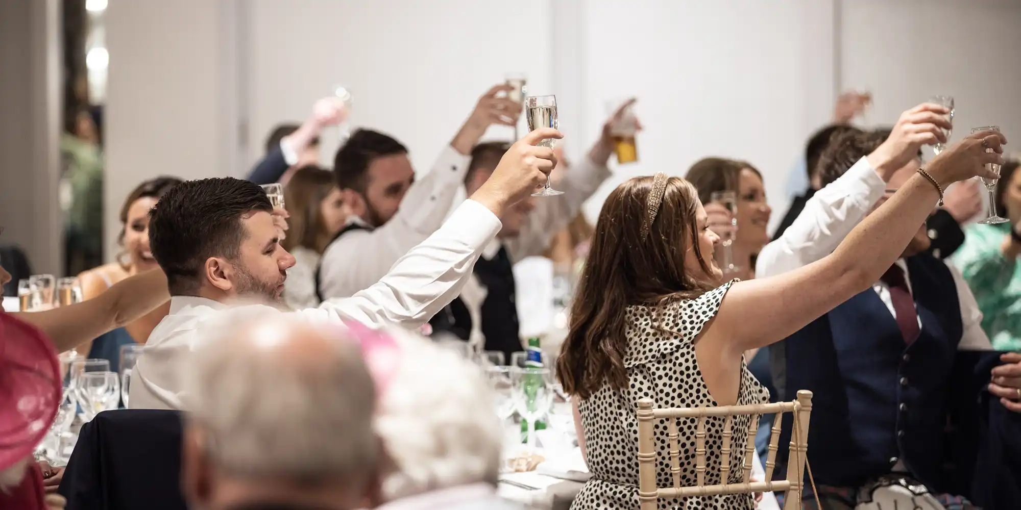 People raising their glasses in a toast at a formal event, with tables set for a meal and guests dressed in formal attire.