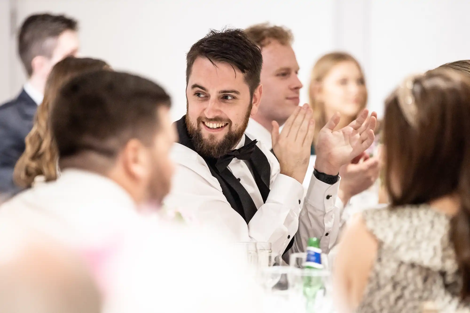 A man in formal attire, wearing a black bow tie, is smiling and clapping at a social event. He is seated among a group of people, some also clapping and smiling.