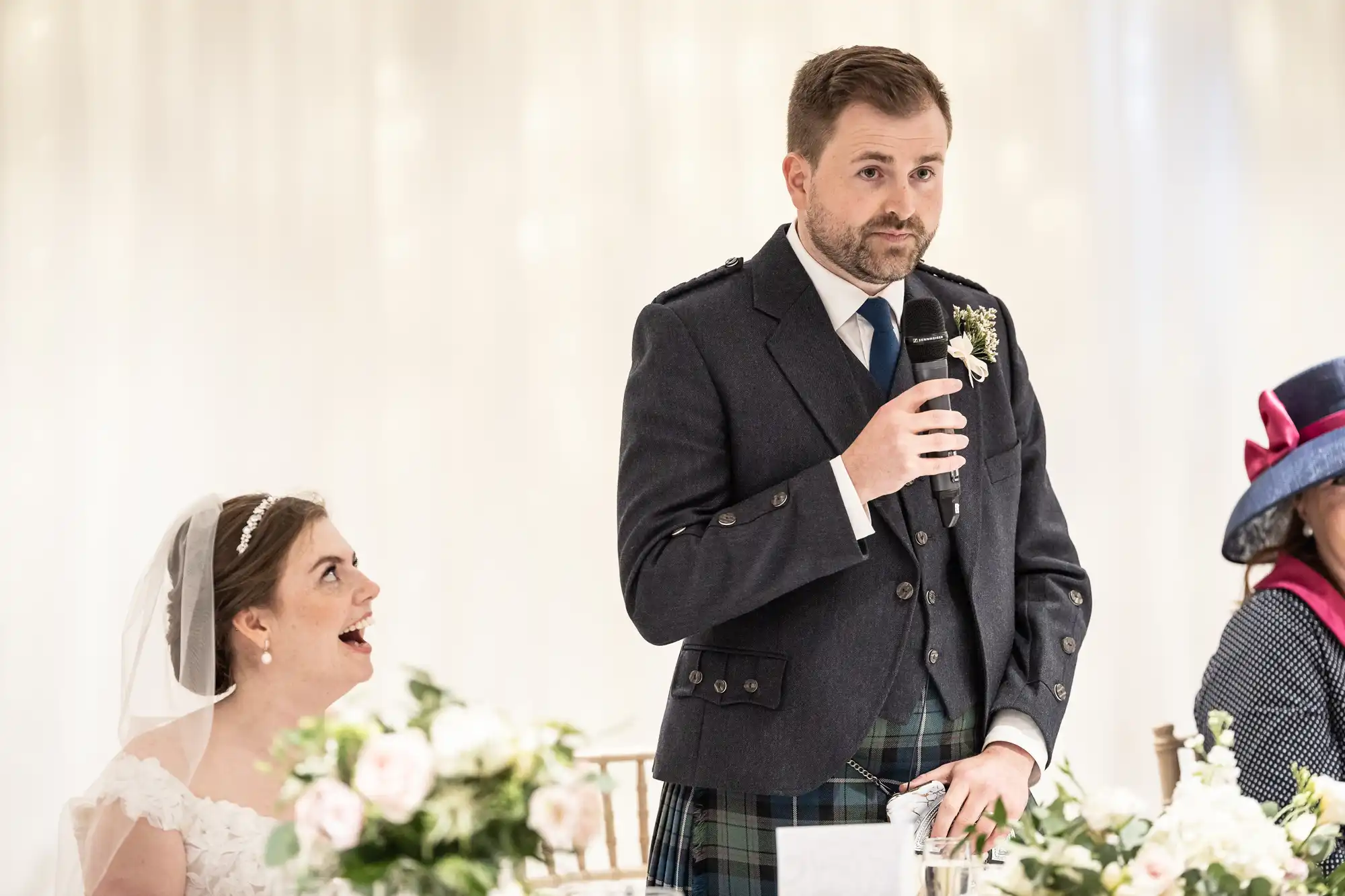 A man in a suit and kilt speaks into a microphone at a formal event. A woman in a wedding dress and a person in a hat are seated beside him.