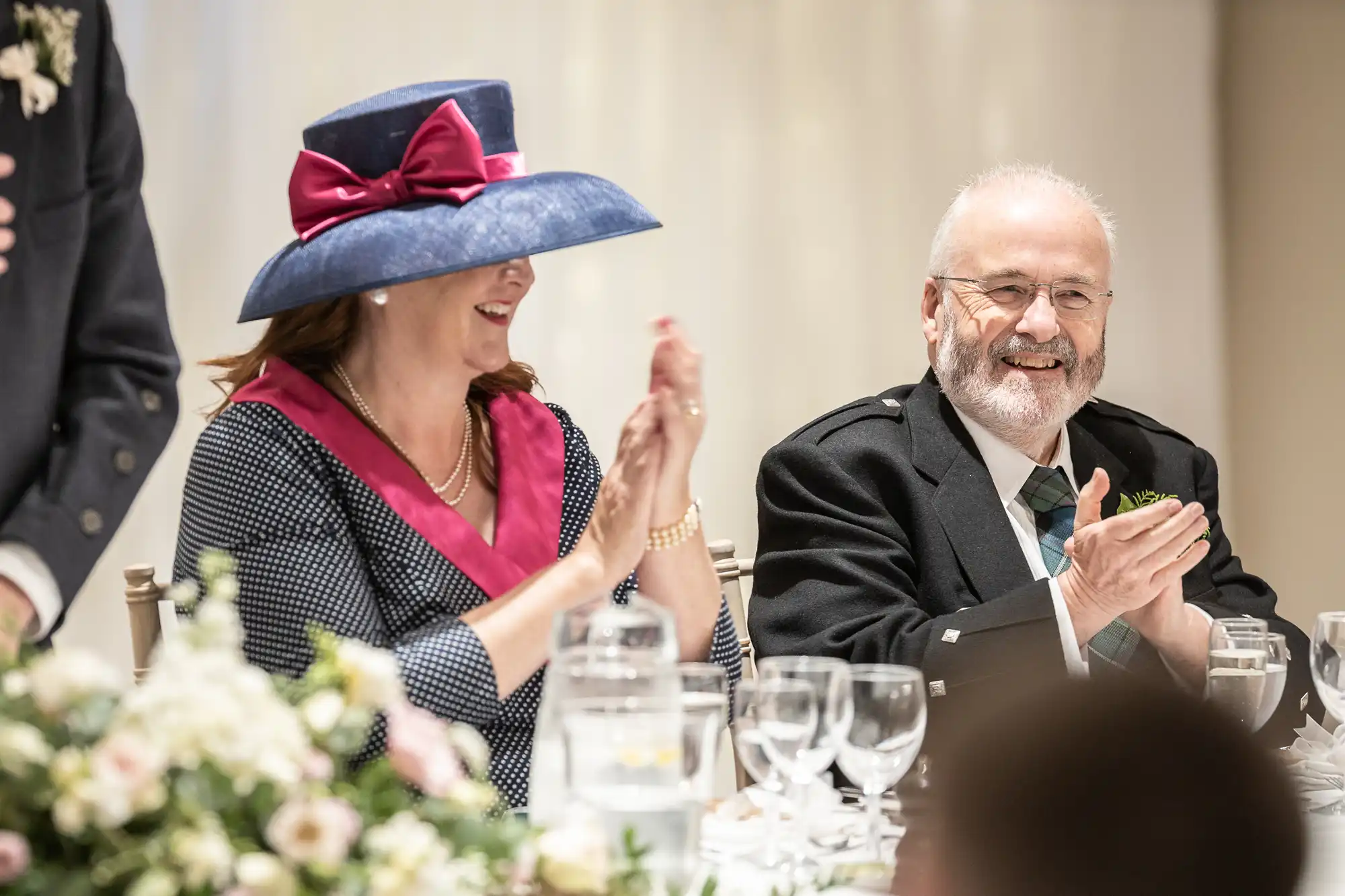 A woman in a blue hat with a red bow and a man in a suit are seated at a table, smiling and clapping, with flowers and glasses in the foreground.