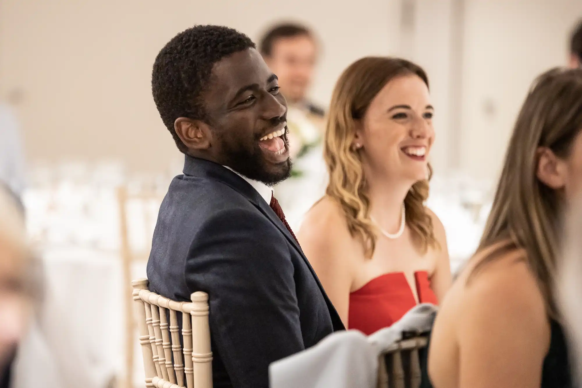 A man and woman sitting next to each other, both laughing and looking to their right, at an indoor event with other people in the background.