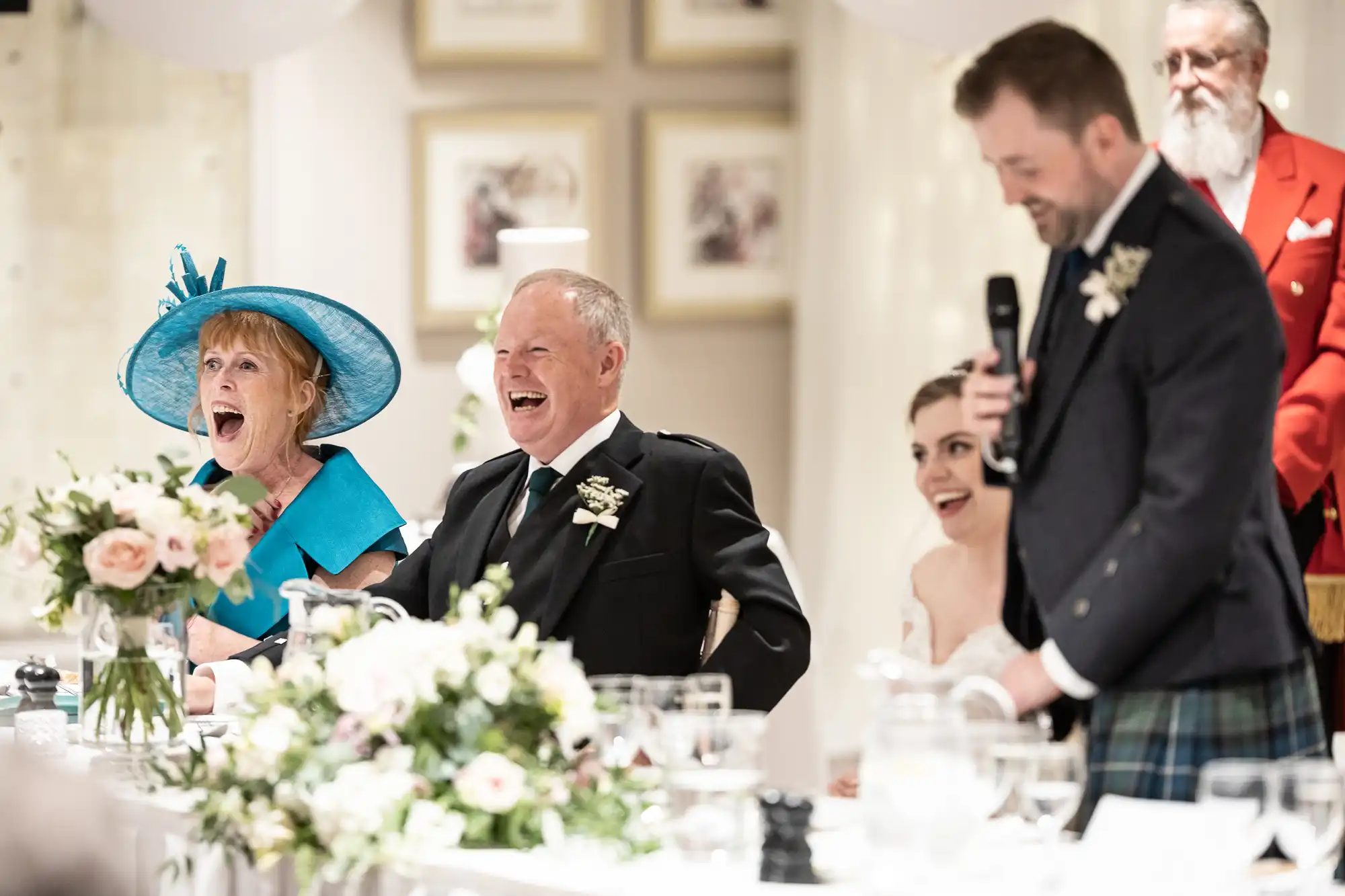 A wedding reception with a speaker addressing the guests. The bride, groom, and two other attendees, one in a blue hat, are seated and laughing. A bouquet and table settings are visible in the foreground.