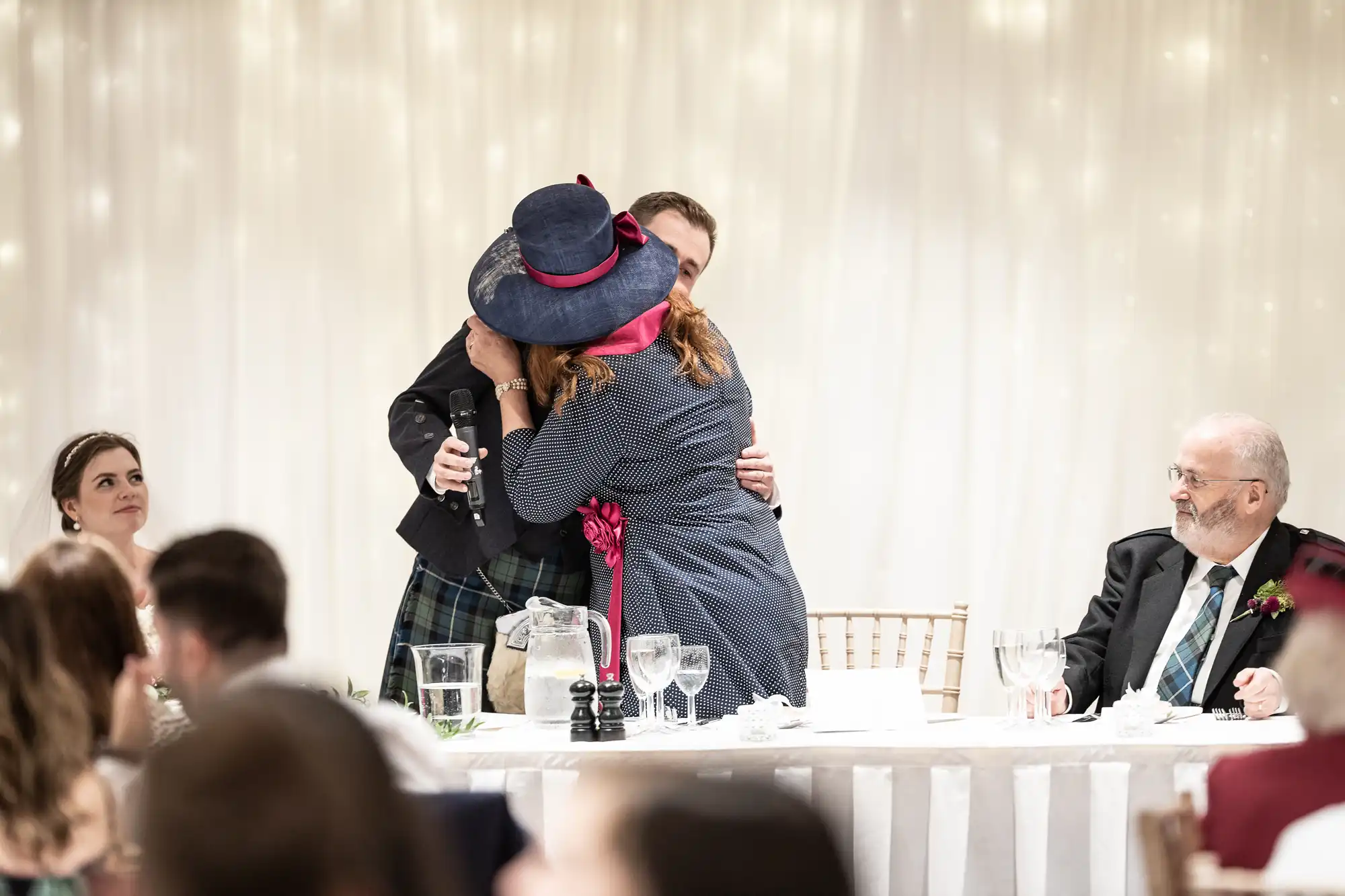 Two people embrace at a wedding reception while others look on from the head table. White tablecloths and twinkling lights create a festive atmosphere.