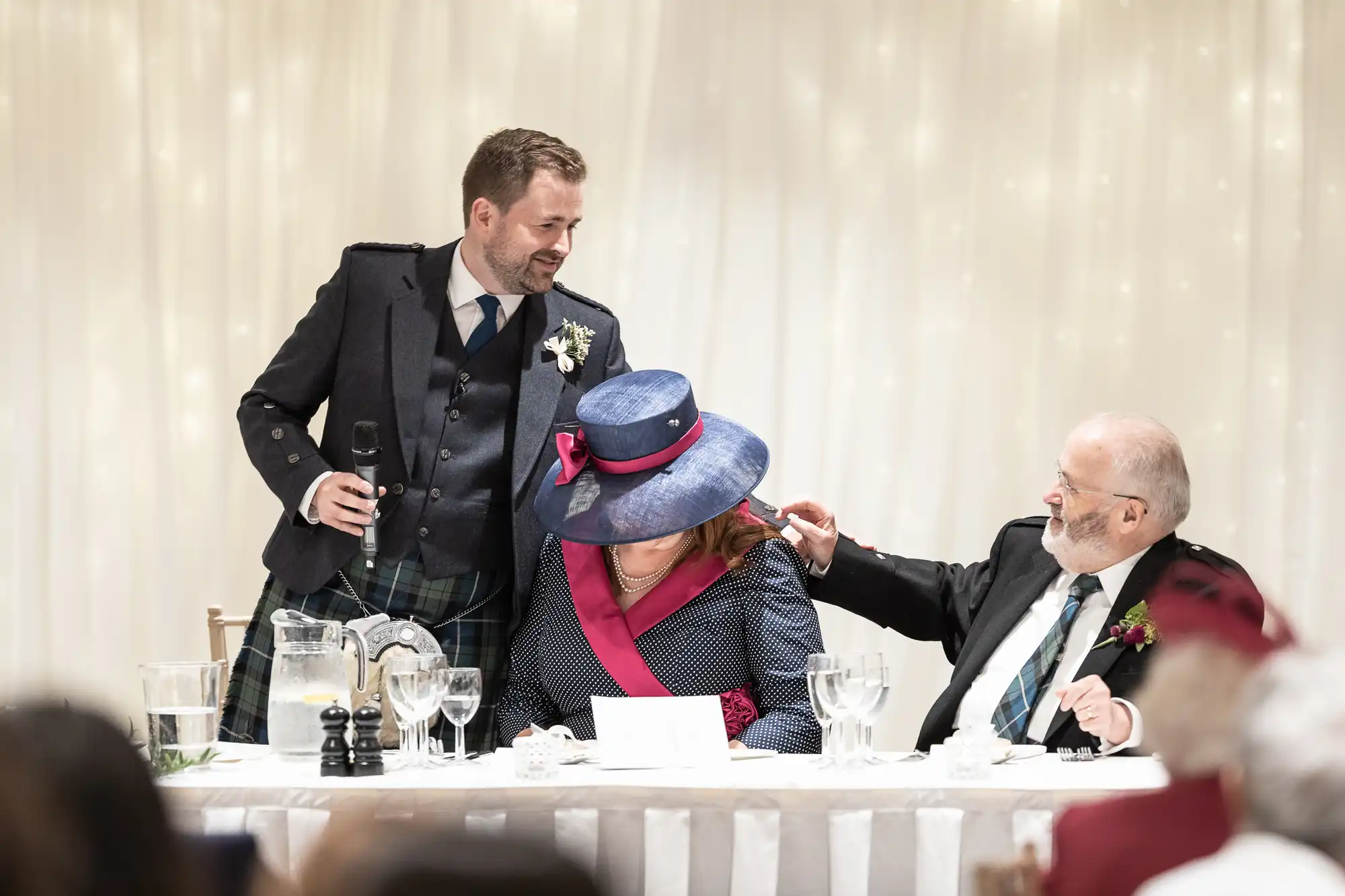 A man in a kilt stands holding a microphone, addressing a seated woman in a blue hat and another man with a beard, both dressed formally, at a decorated event table.