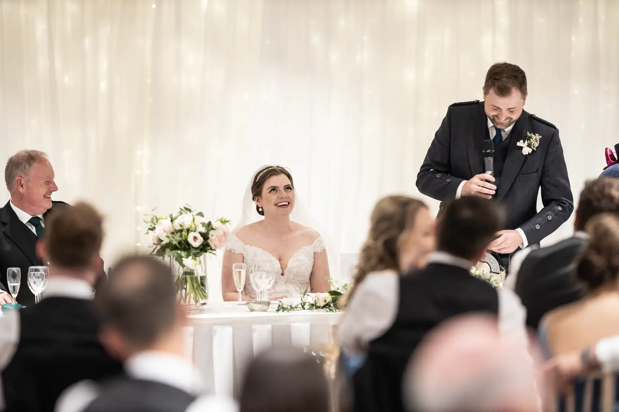 A bride in a white dress and a groom in a dark suit are seated at a table. The groom is speaking into a microphone while the bride looks up at him, smiling. Guests are seated around them.