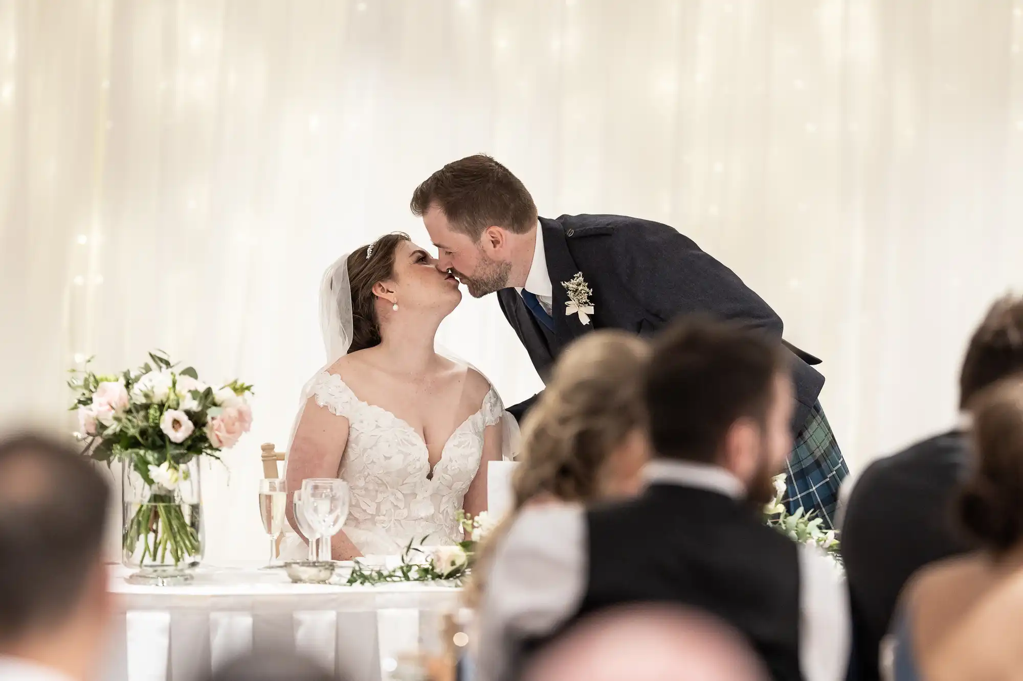 A couple in wedding attire share a kiss at their reception. The bride sits at a table adorned with flowers while the groom leans over her. Guests seated with backs to the camera observe.