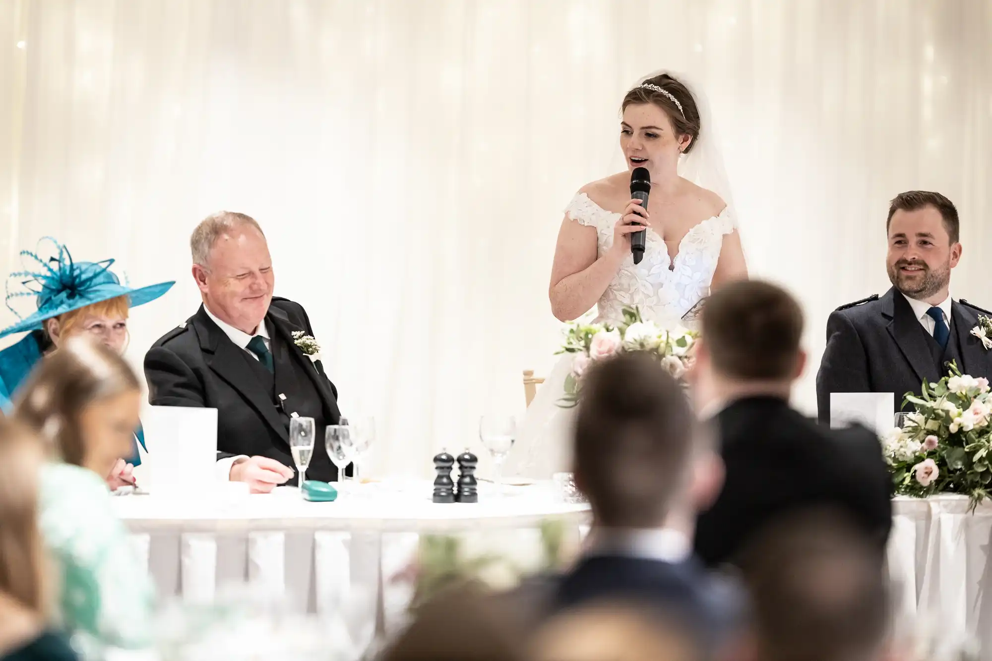 A bride holding a microphone gives a speech at a wedding reception with guests seated at a table in front of her.