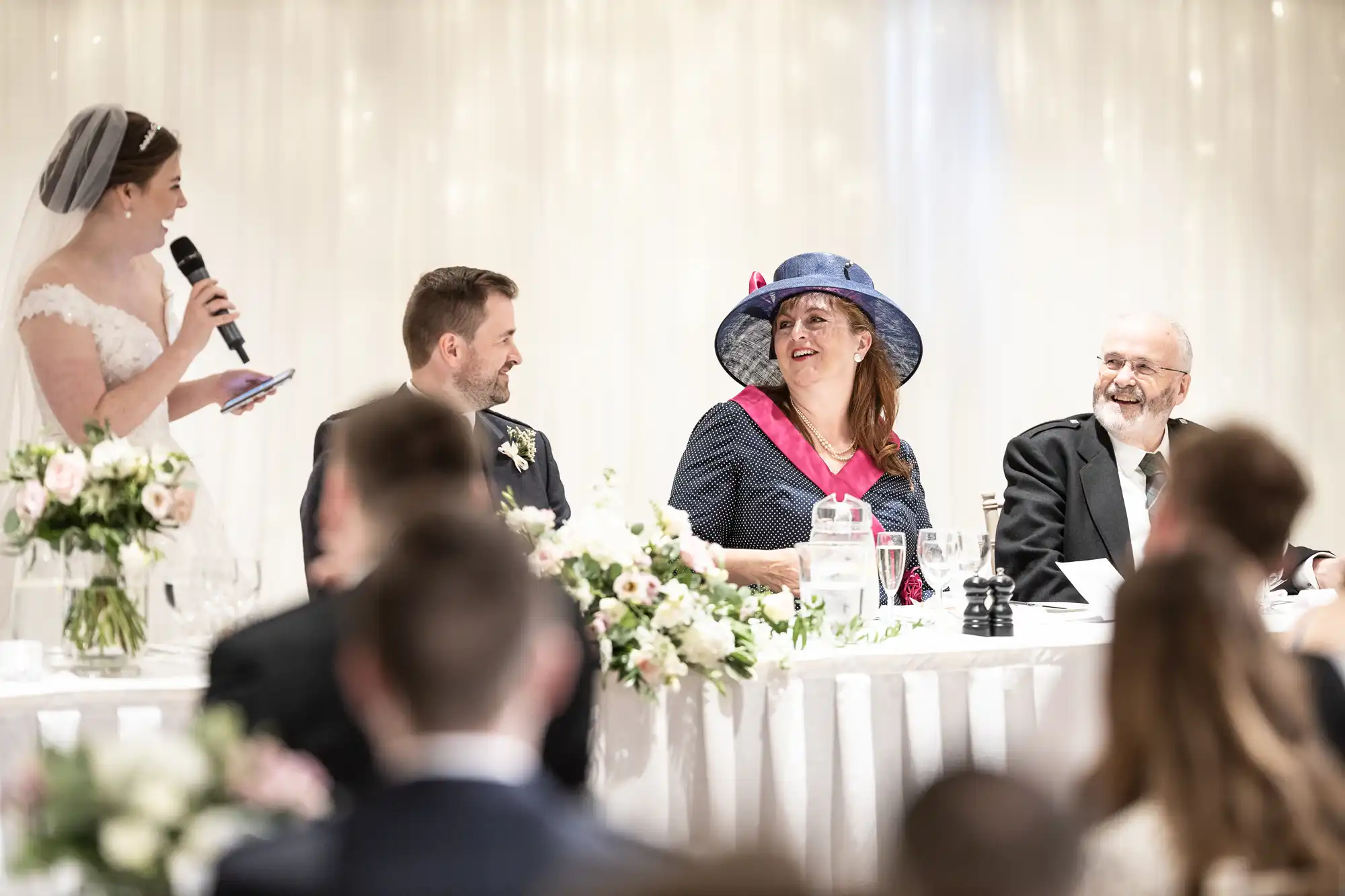 A bride speaks into a microphone at a wedding reception while three people sitting at a long table listen and smile. The table is decorated with flowers and the background is draped with white fabric.