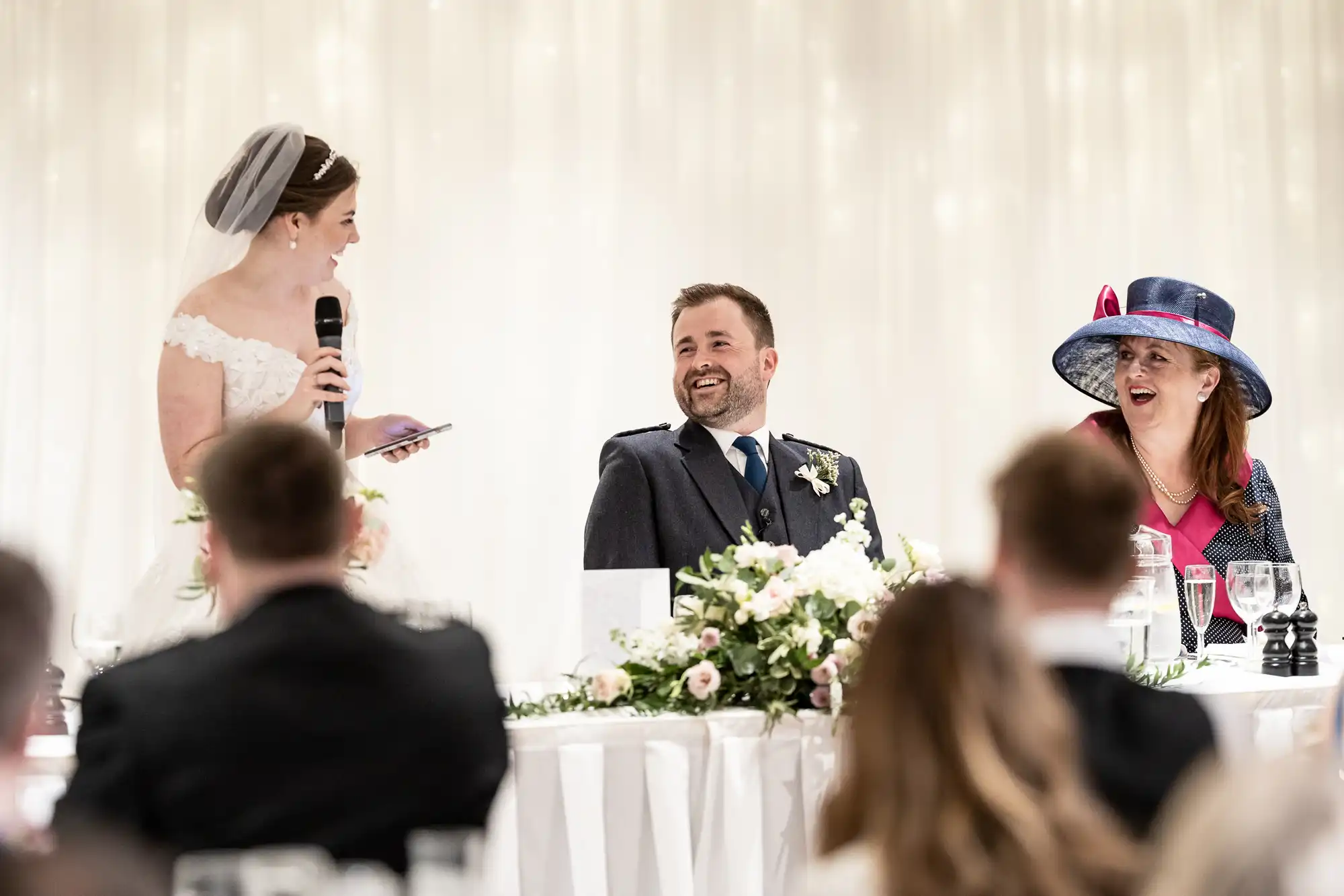 A bride speaks into a microphone while a smiling groom and a woman in a blue hat with a pink ribbon listen, seated at a wedding reception table decorated with flowers and glasses.