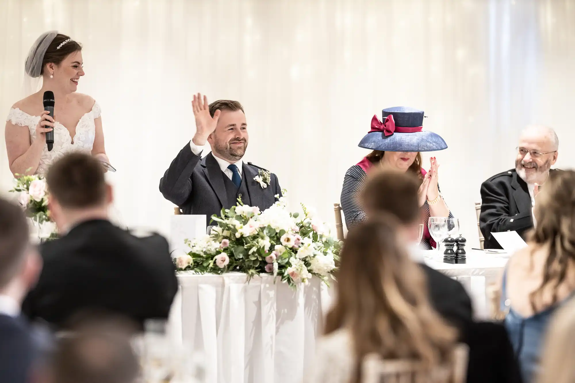 A bride stands with a microphone, while a groom at a table waves. Next to him, a woman in a large hat claps, and an older man sits smiling. They are at a wedding reception with flower arrangements.