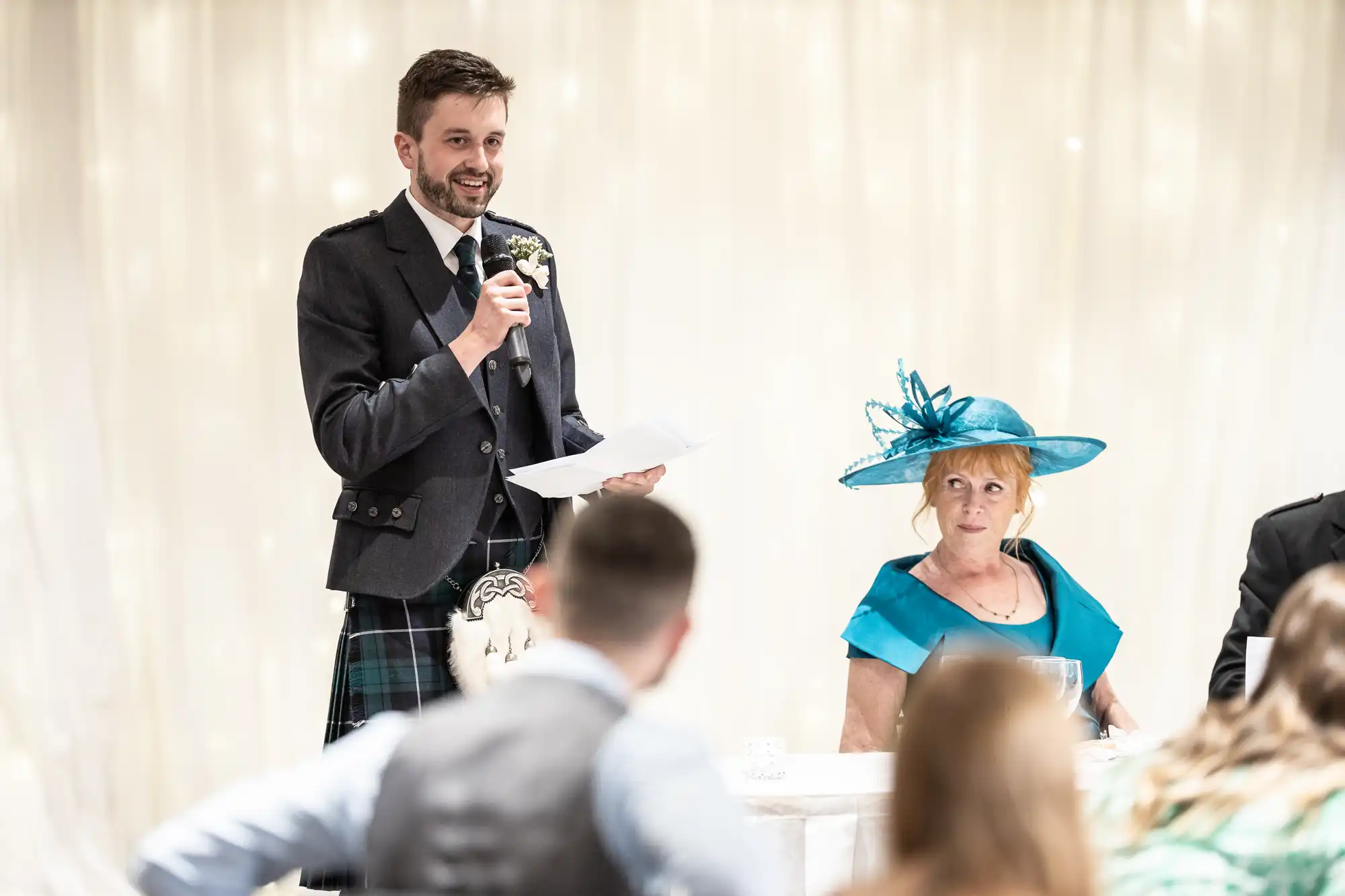 A man in a kilt speaks into a microphone while holding a paper, standing beside a seated woman in a teal dress and hat at an event.