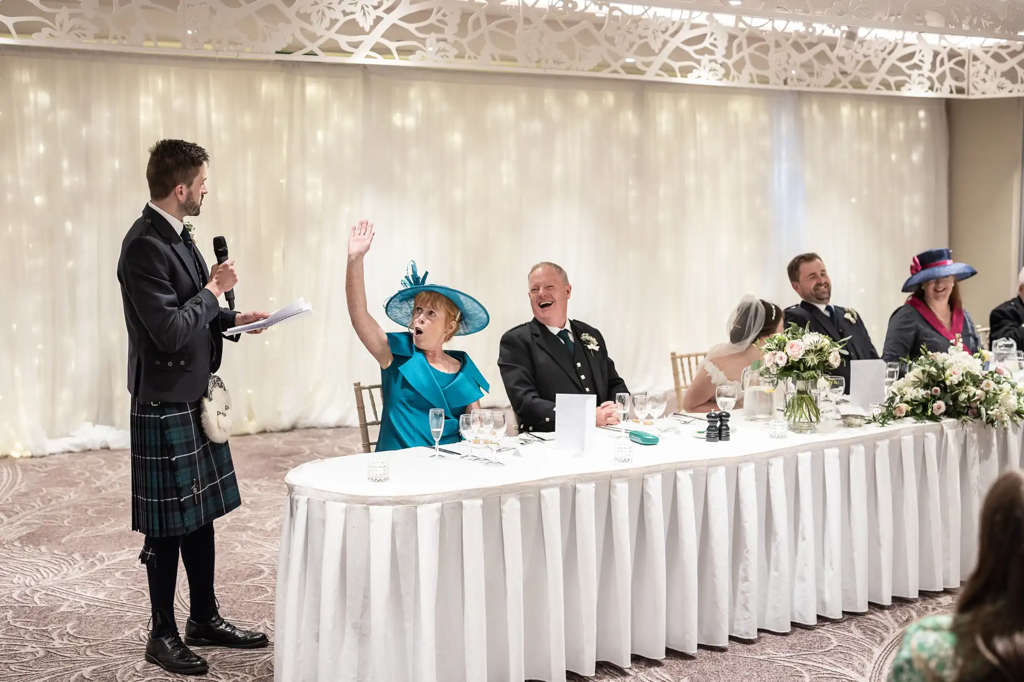 A woman in a bright blue dress raises her hand while seated at a long table with other guests. A man in a kilt speaks into a microphone, standing to her left. The table is decorated with flowers.