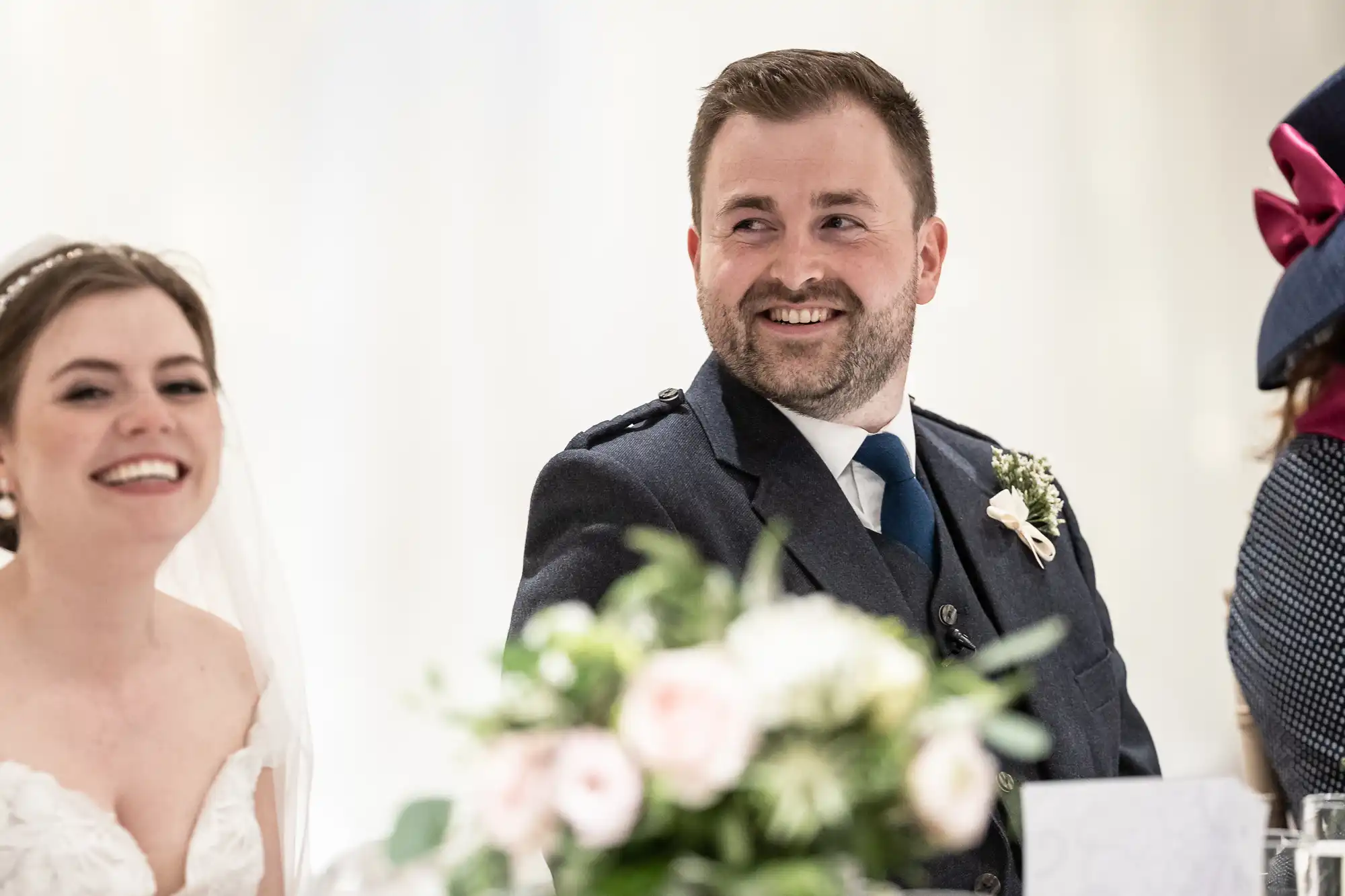 A bride and groom smiling at a wedding reception table, with floral arrangements in the foreground.