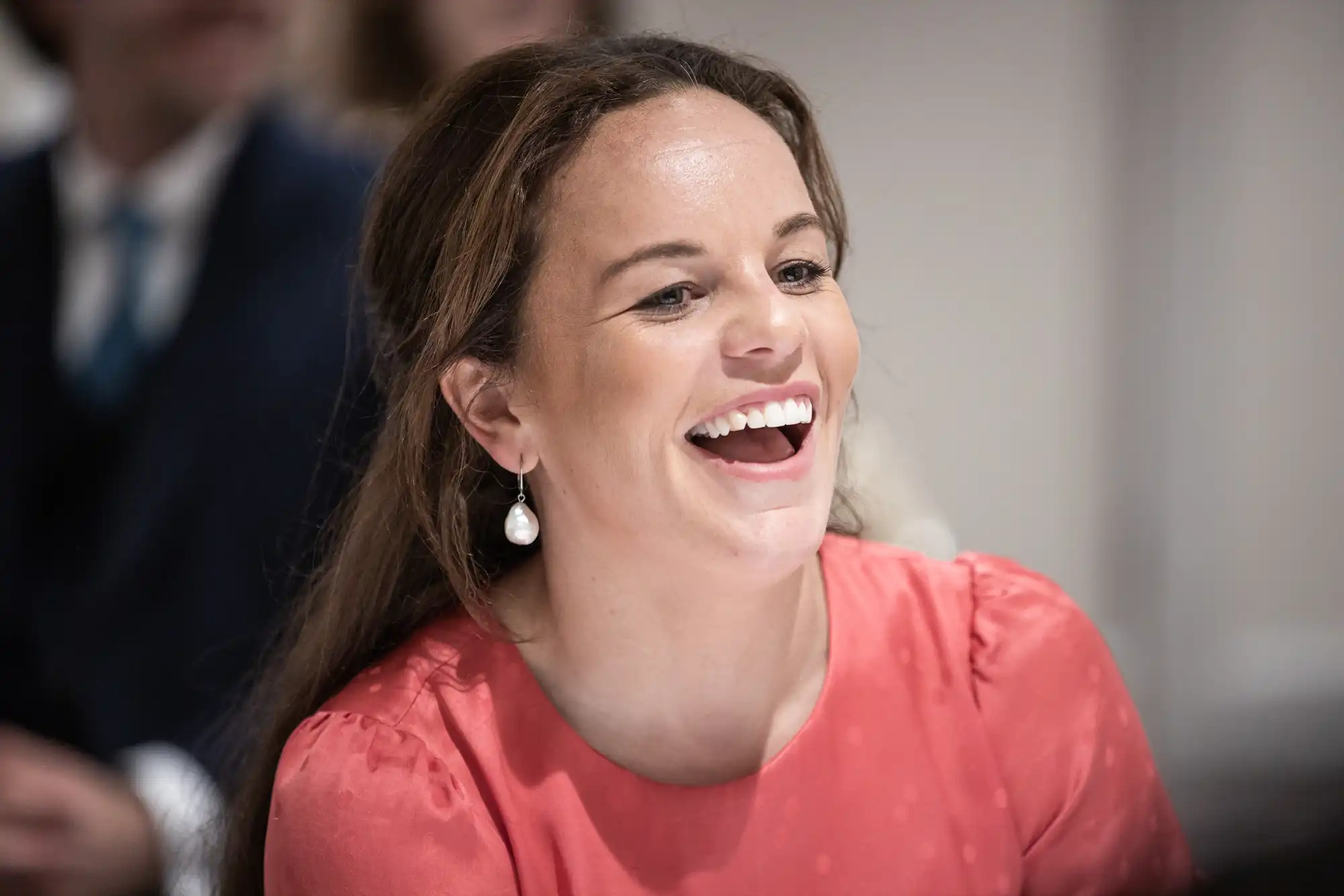 A woman with long hair, wearing a pink top and pearl earrings, is smiling and looking slightly to her left.