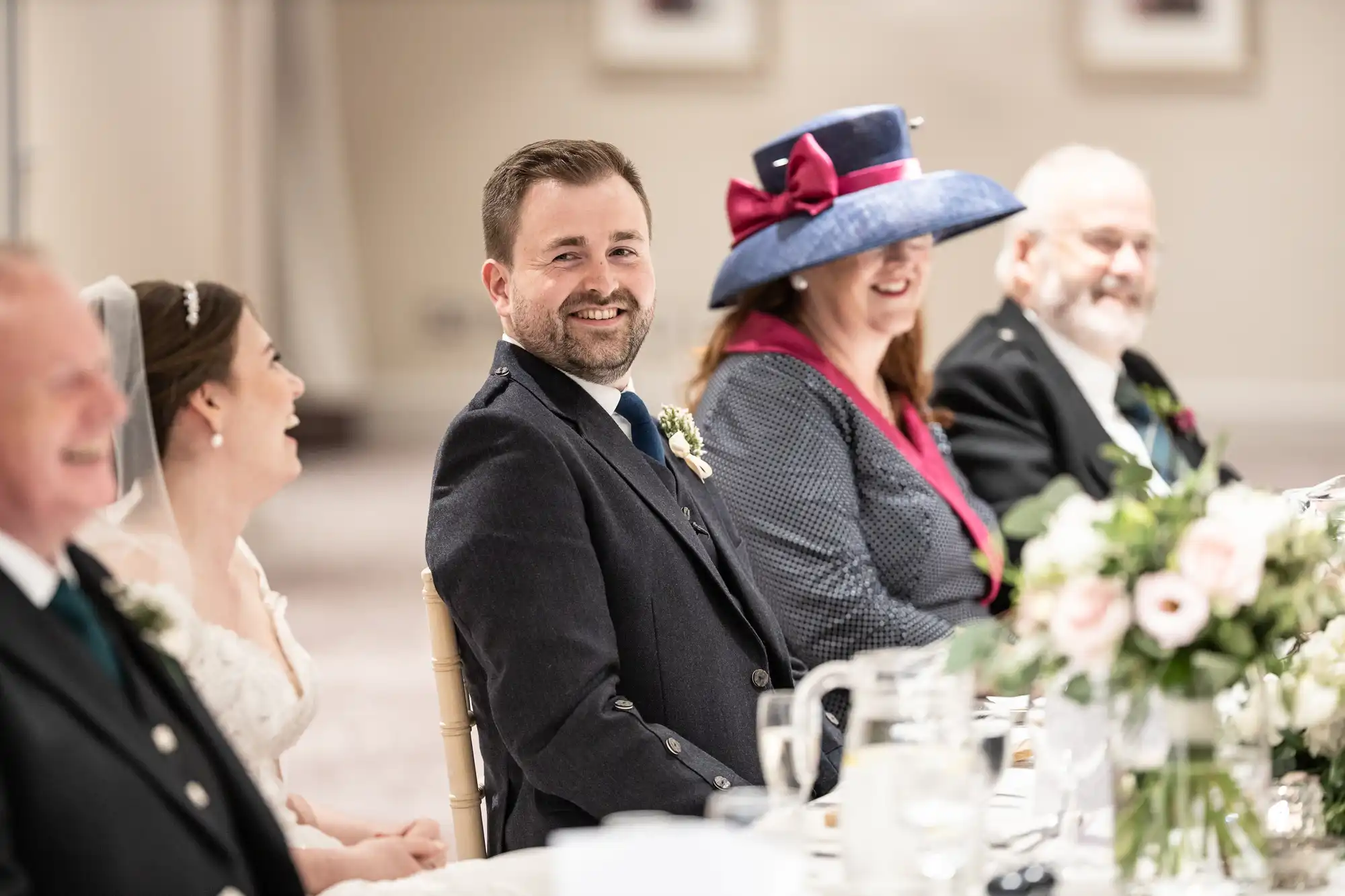 A group of people in formal attire, including a bride and groom, sit at a table during a wedding reception, smiling and laughing.