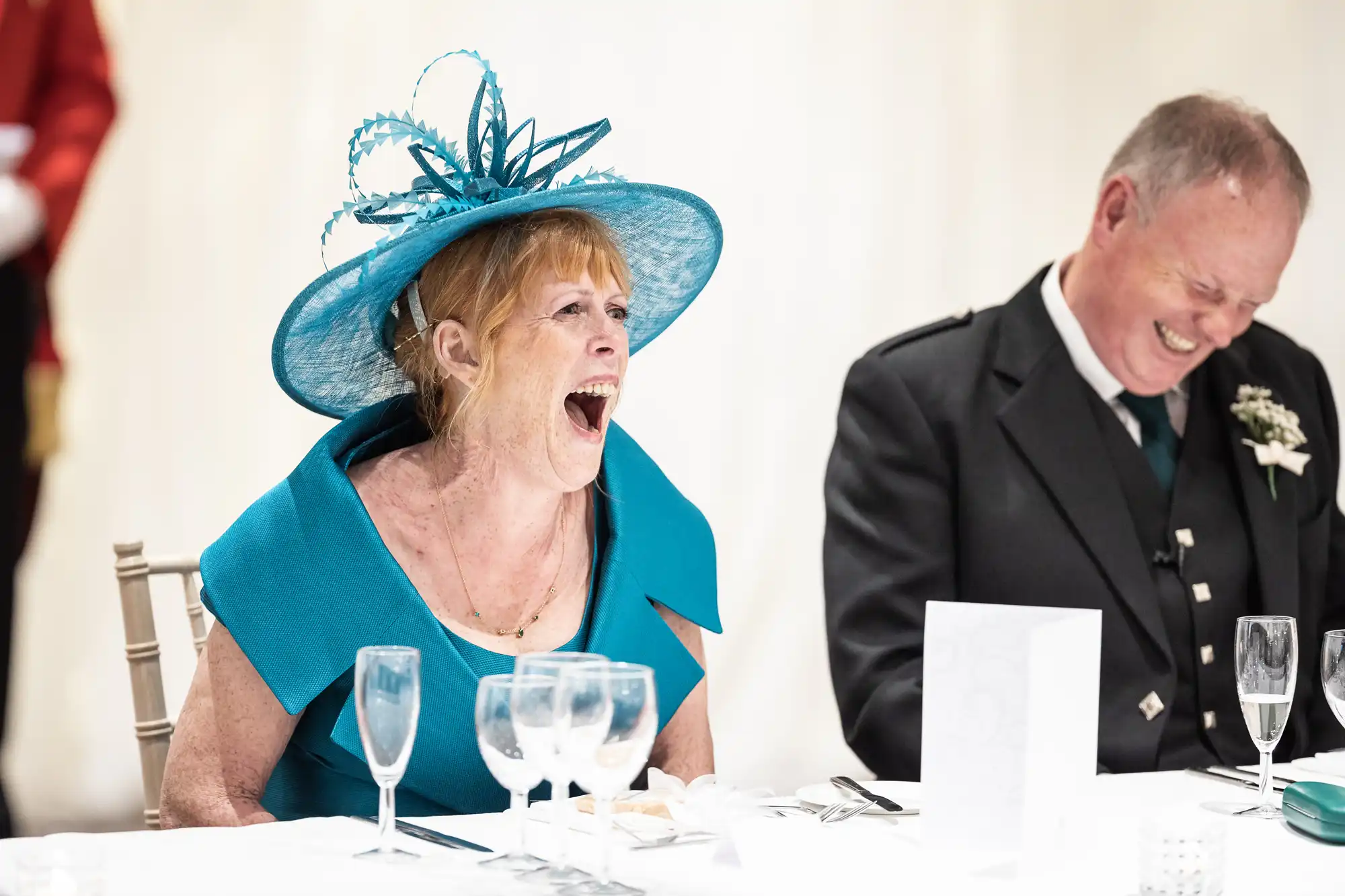 A woman in a blue hat and dress laughs heartily while sitting at a table next to a man in formal attire who is also laughing.