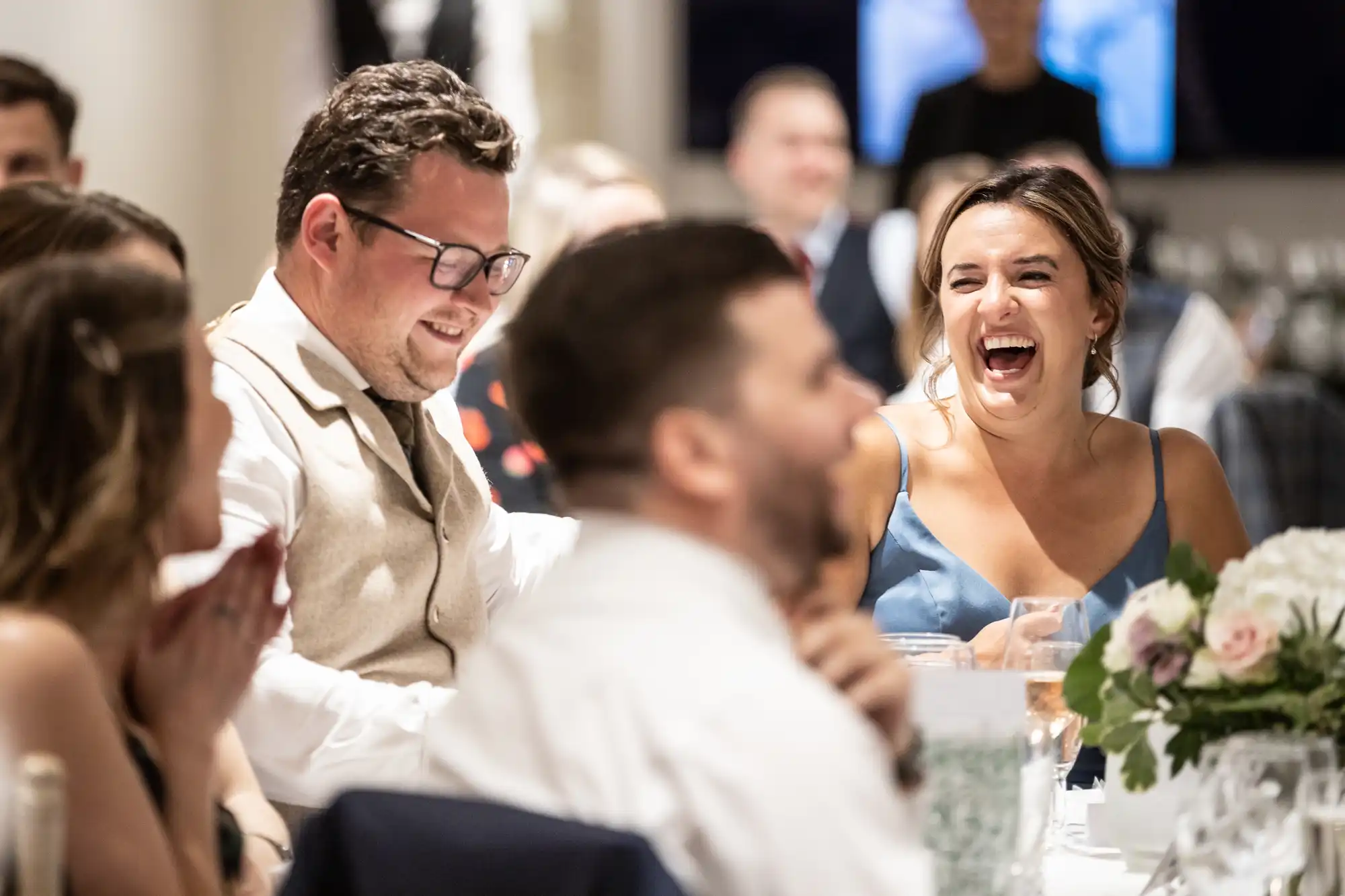 A group of people, dressed in formal attire, are seated at a table and laughing. A woman in a blue dress and a man in a beige vest are seen prominently. Flowers are visible on the table.